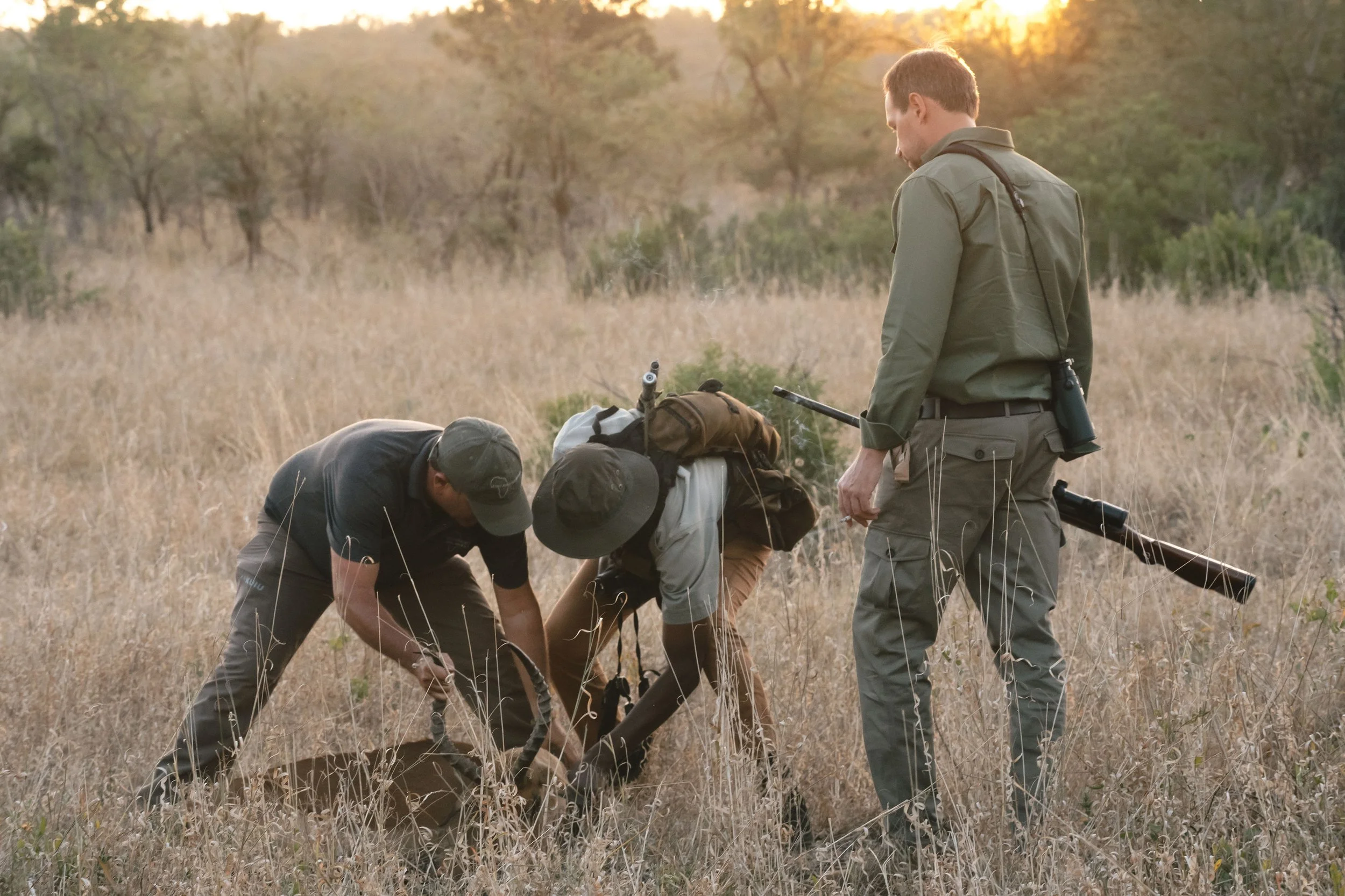 Three hunters in khaki and black clothing, with one wearing a backpack and one holding a rifle, are handling a dog in a dry grassy field at sunset.