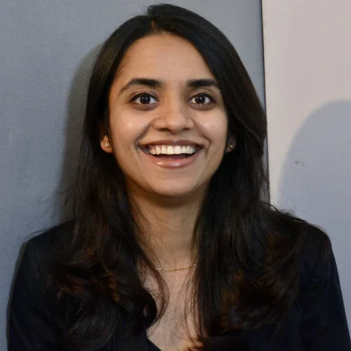 A young woman with long dark hair smiling at the camera, wearing a black blazer against a plain background.