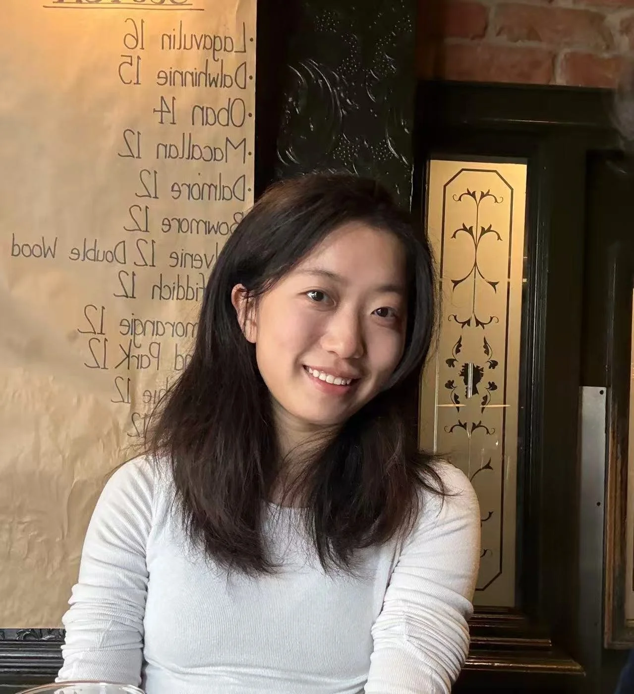 A young woman with shoulder-length dark hair smiling in front of a menu board and decorative wall.