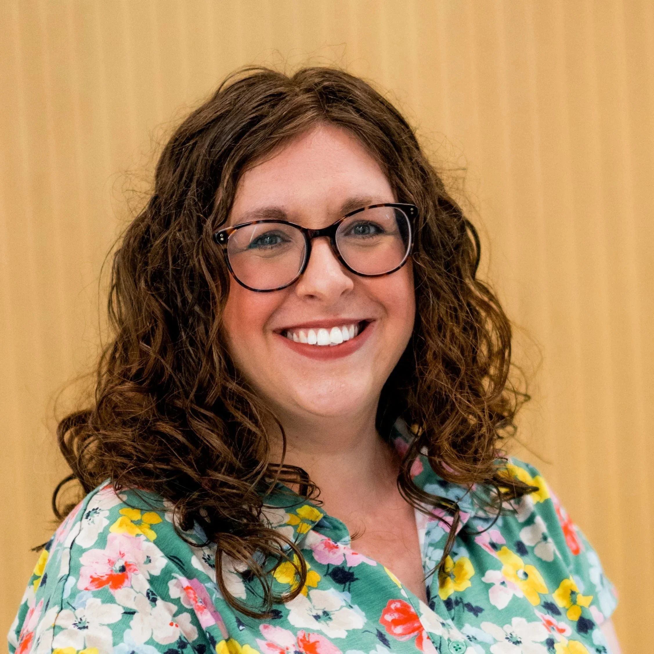 A woman with curly brown hair, glasses, and a bright smile, wearing a colorful floral shirt, standing against a wooden background.