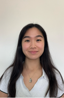 A young woman with long dark hair, smiling, wearing a white top and a necklace, standing against a plain light-colored background.