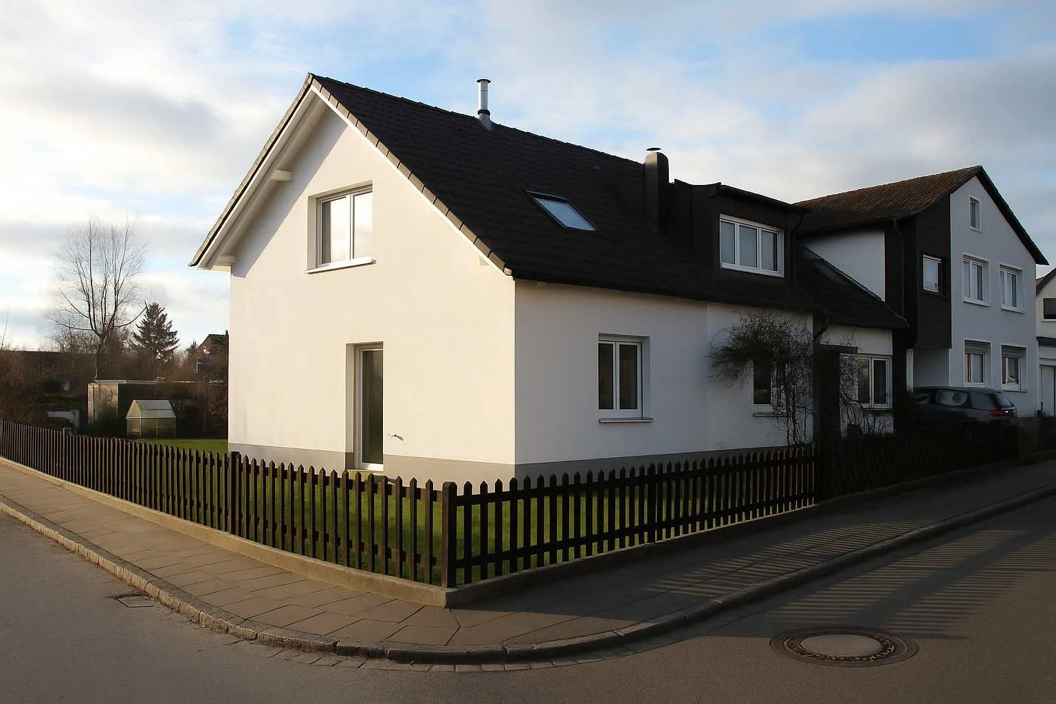 Ein modern gehaltenes, weißes Einfamilienhaus mit dunklem Satteldach, umgeben von einem kleinen Garten mit Zaun, an einer ruhigen Nebenstraße im Hellen an einem sonnigen Tag.
