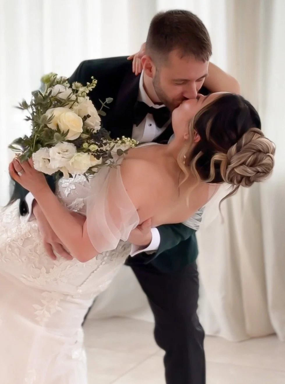 A black-and-white photograph of a newlywed couple kissing outdoors, surrounded by wedding guests. The bride, holding a large bouquet of flowers, is wearing an off-the-shoulder wedding dress and veil, while the groom is in a tuxedo with a bow tie.