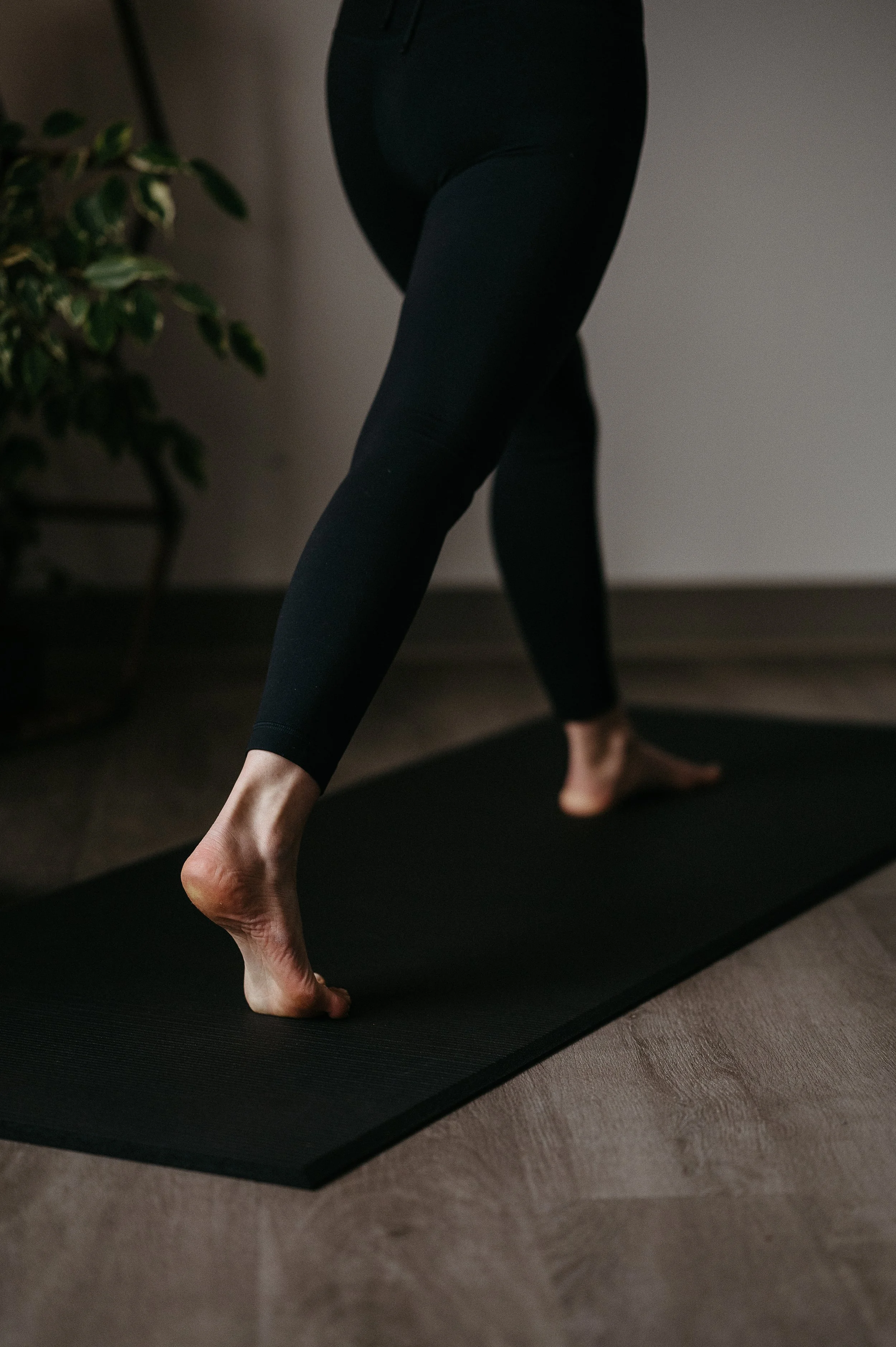 A person practicing yoga on a black mat indoors, wearing black leggings, with only the lower body visible. One foot is on the mat, and the other is raised with toes pointed downward, in a stretching pose. A green plant is partially visible in the background.