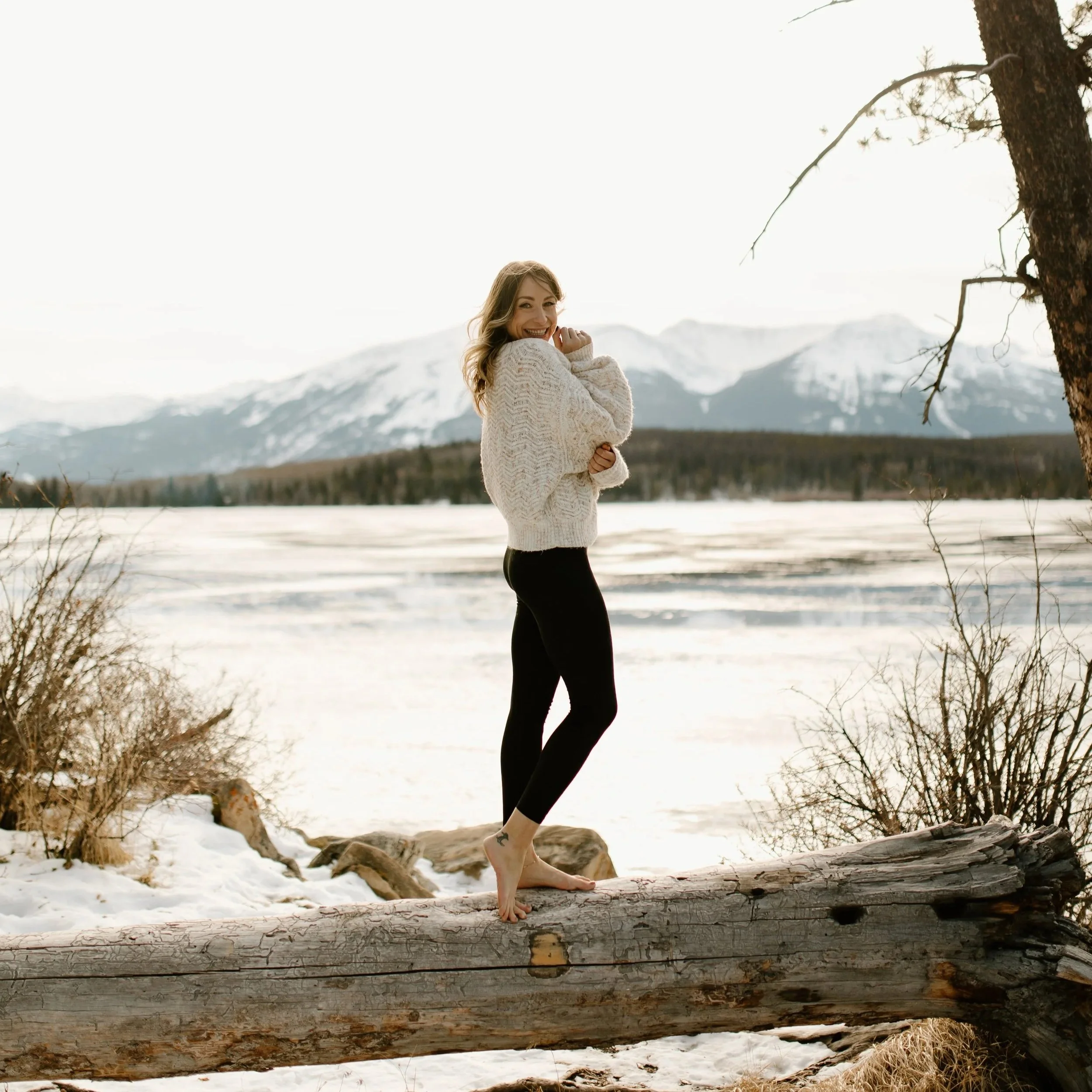 A woman smiling and standing on a fallen tree trunk in a snowy landscape with mountains and a tree in the background.
