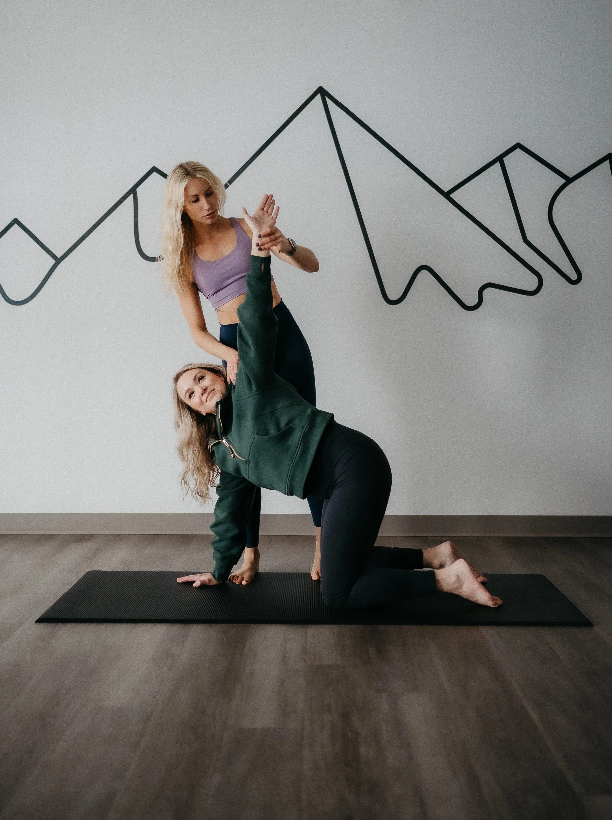 Two women practicing yoga indoors on a black mat, with a wall featuring a black line drawing of a mountain range in the background.