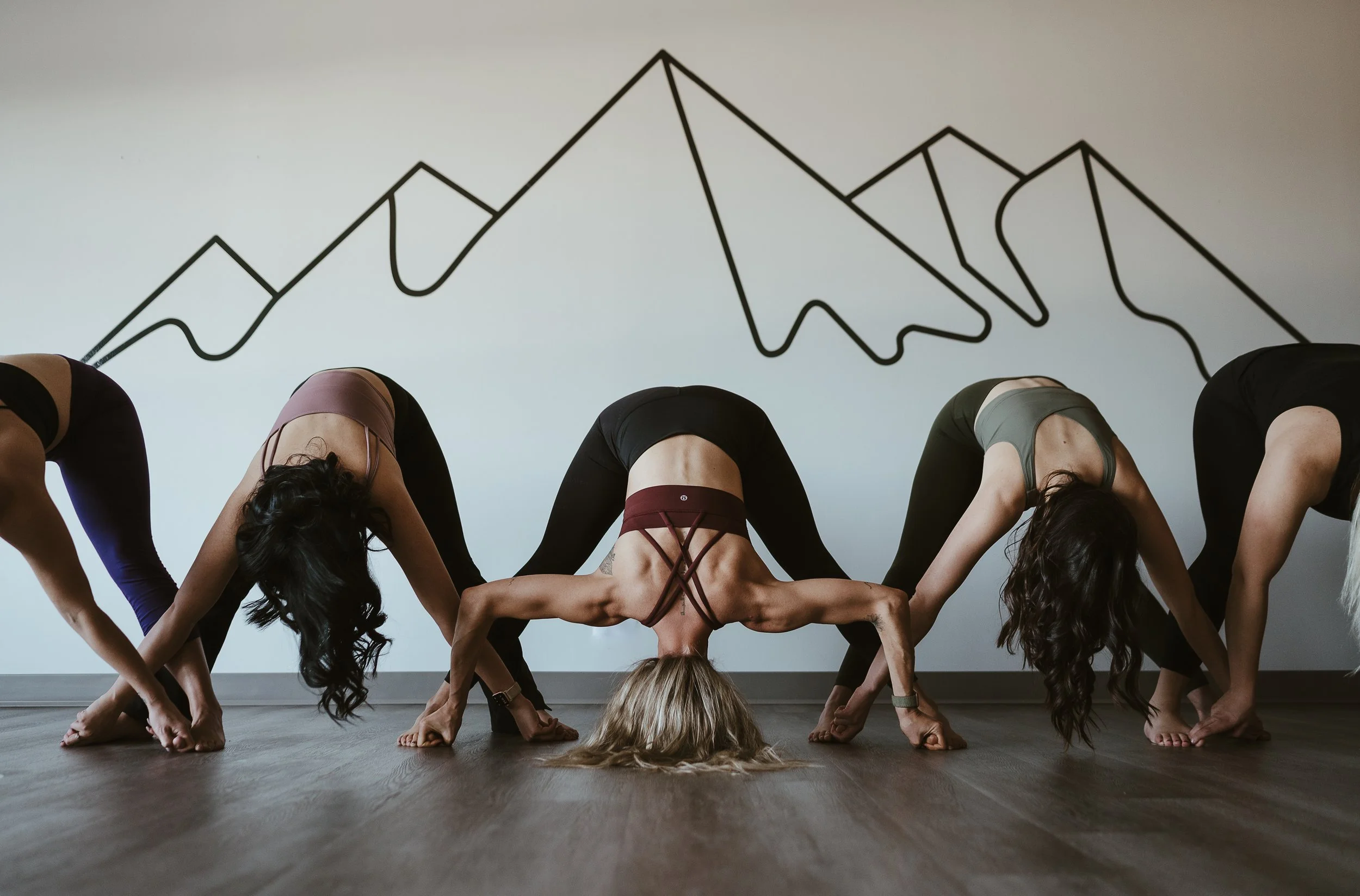Group of women practicing yoga indoors, performing a headstand with hands on the floor and legs bent over their head, in front of a wall with mountain line art.