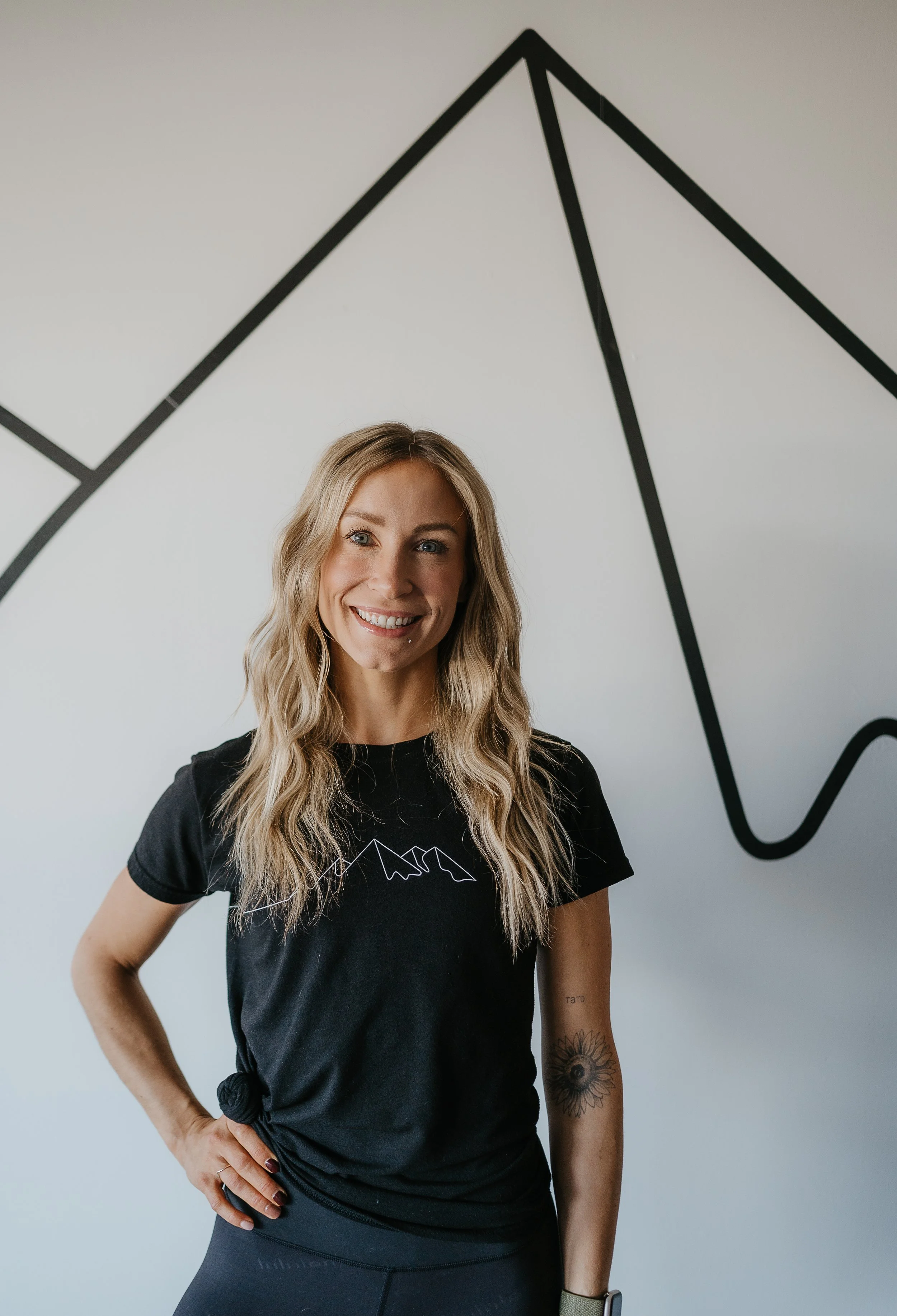 A woman with long blonde wavy hair, wearing a black T-shirt with a mountain outline, standing in front of a white wall with black line art of a mountain and a line graph.