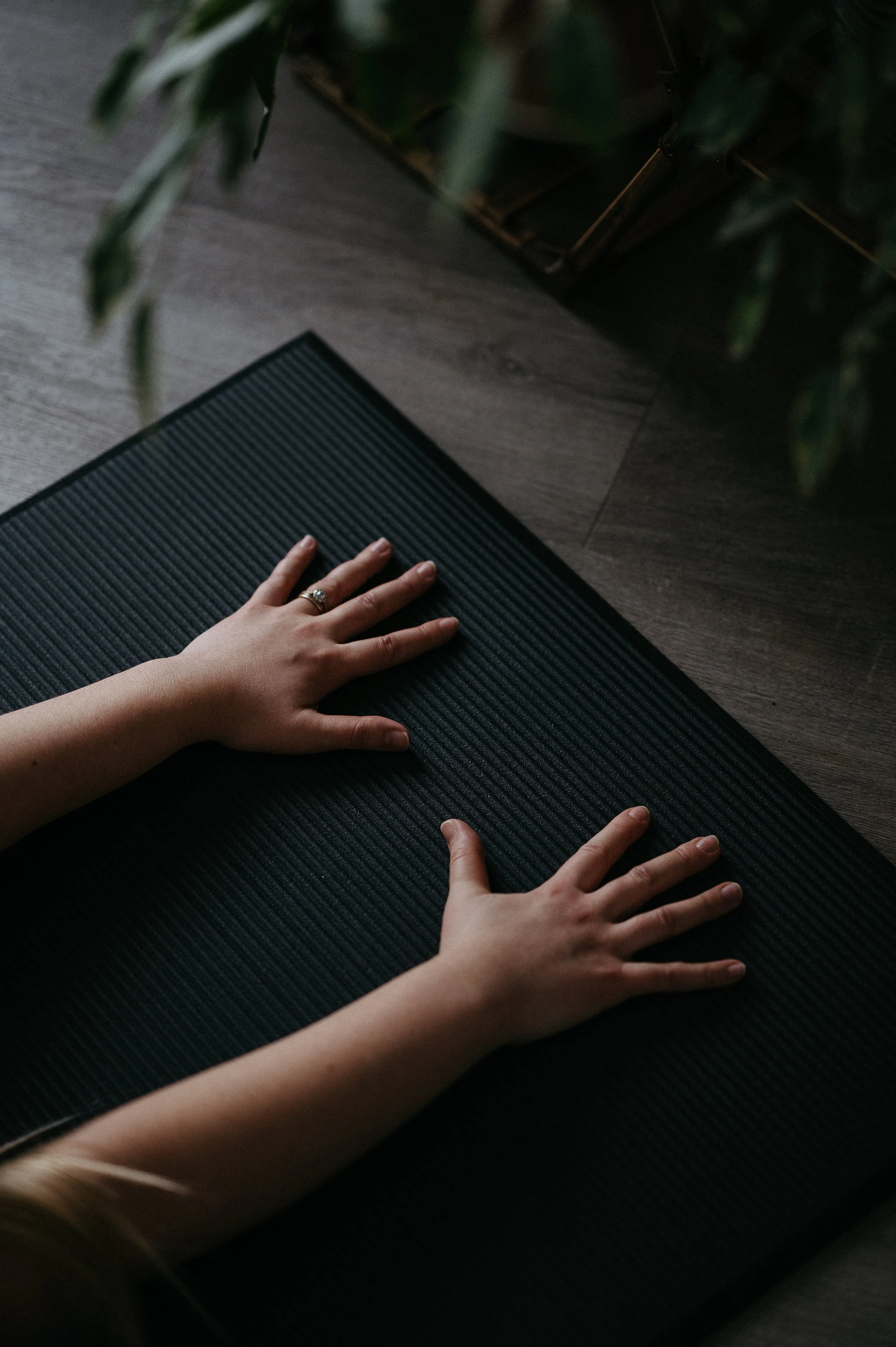 Two hands resting on a black textured yoga mat on a wooden floor, with some green foliage at the top of the image.