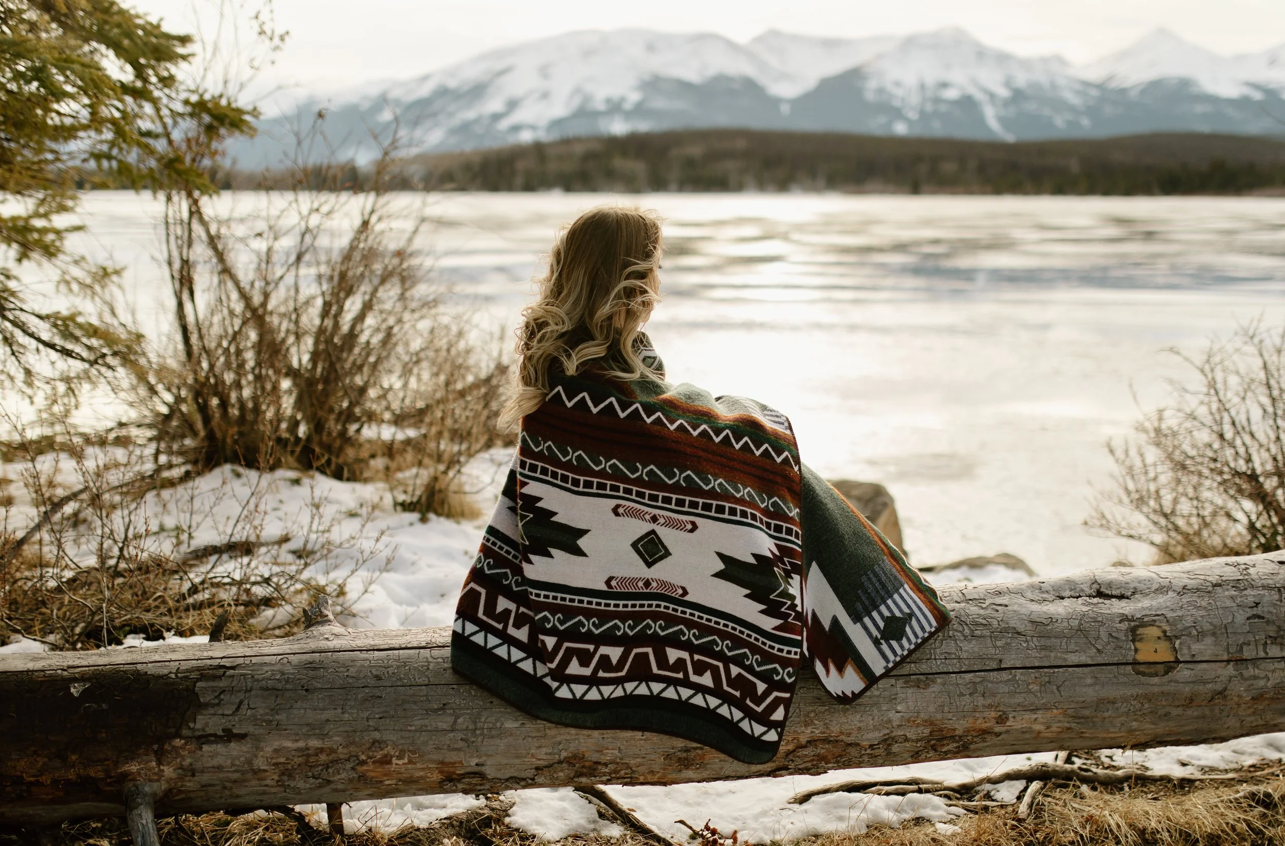 A woman with long blonde hair wrapped in a colorful patterned blanket sitting on a fallen log by a snowy lakeshore, with snow-capped mountains in the background.