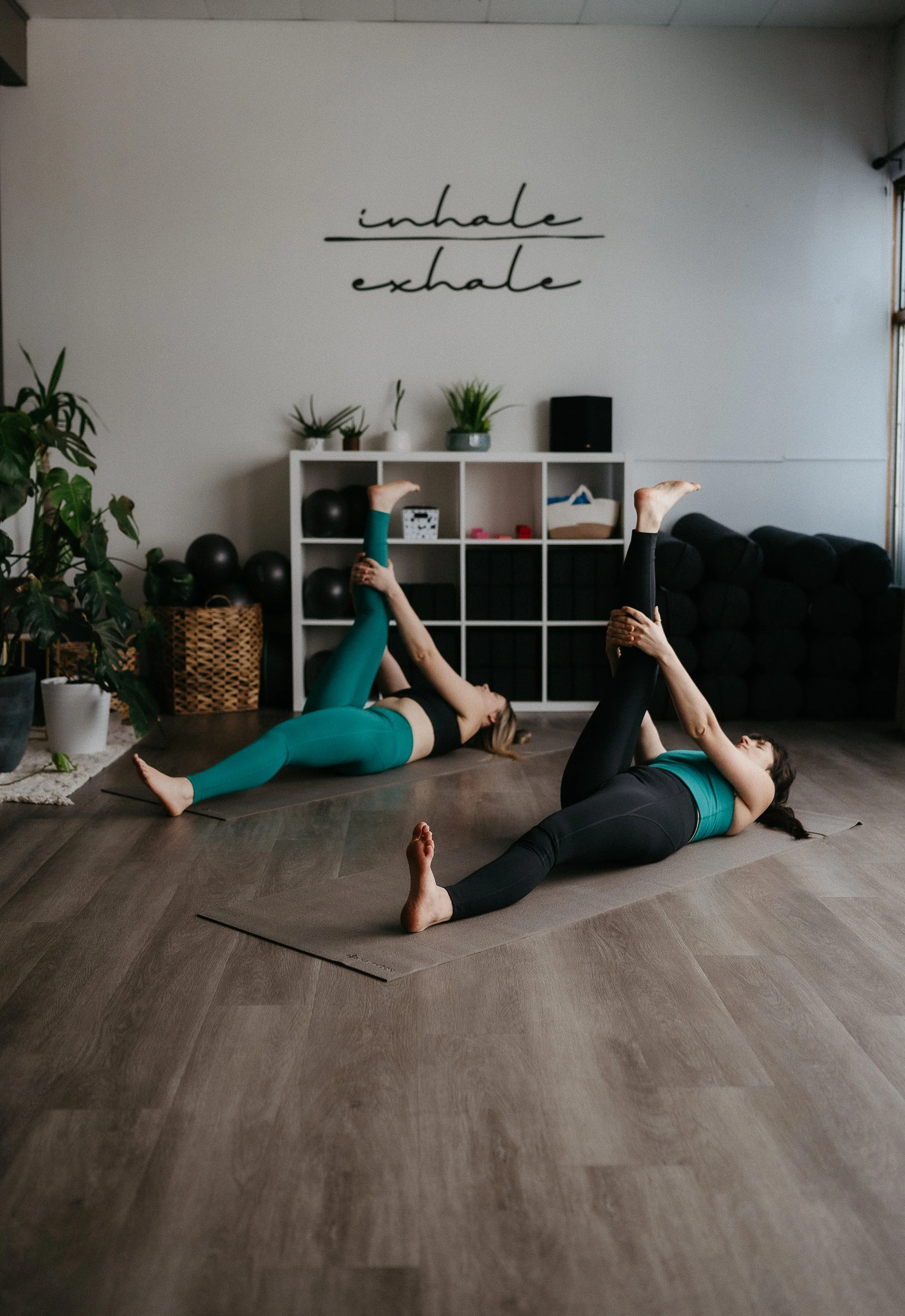 Two women practicing yoga on mats in a yoga studio, with plants and a shelf in the background, and a motivational quote on the wall that says 'inhale' and 'exhale'.
