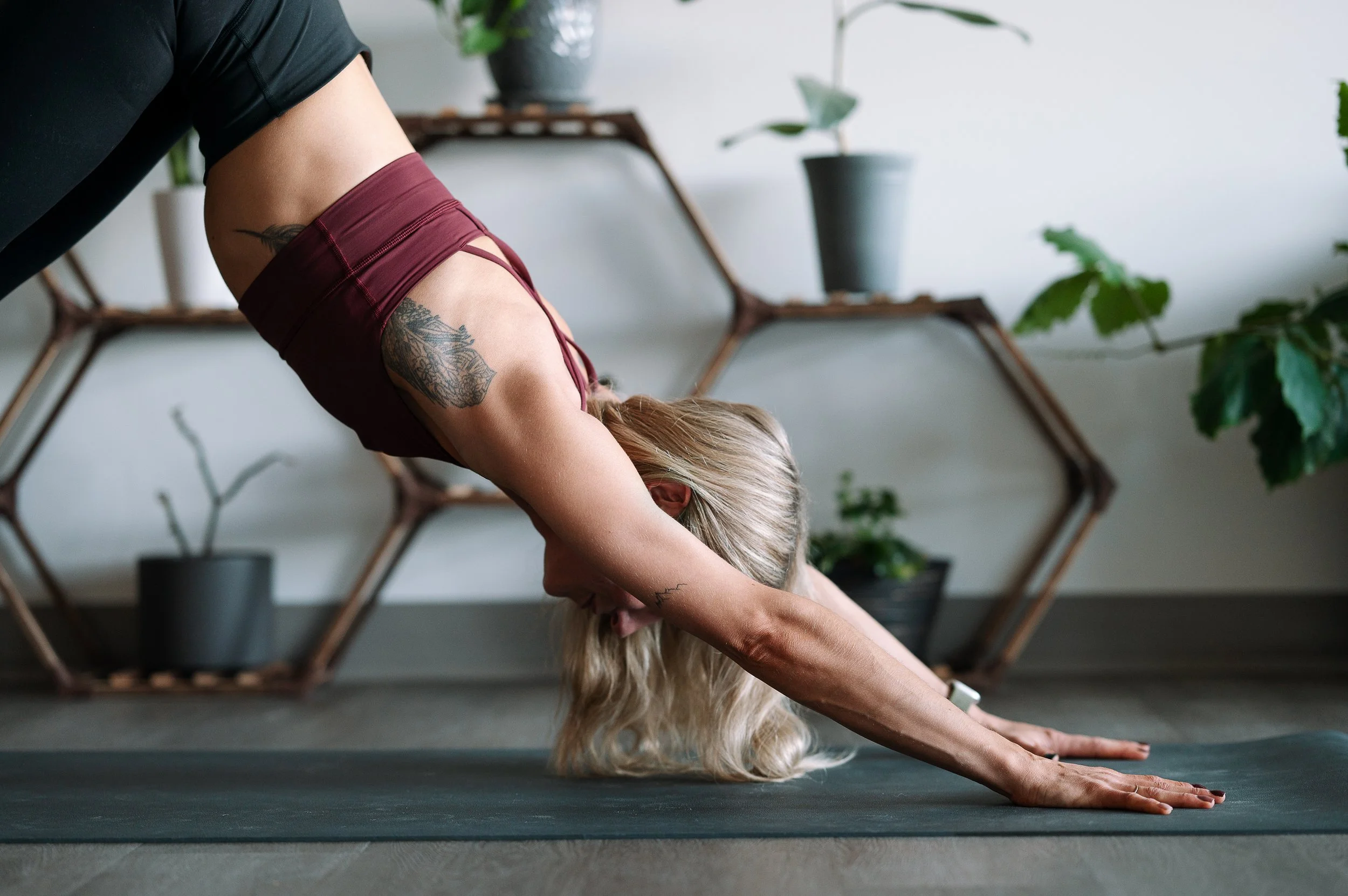 A woman practicing yoga in a downward-facing dog on a dark grey yoga mat in a room with potted plants and wooden shelves.