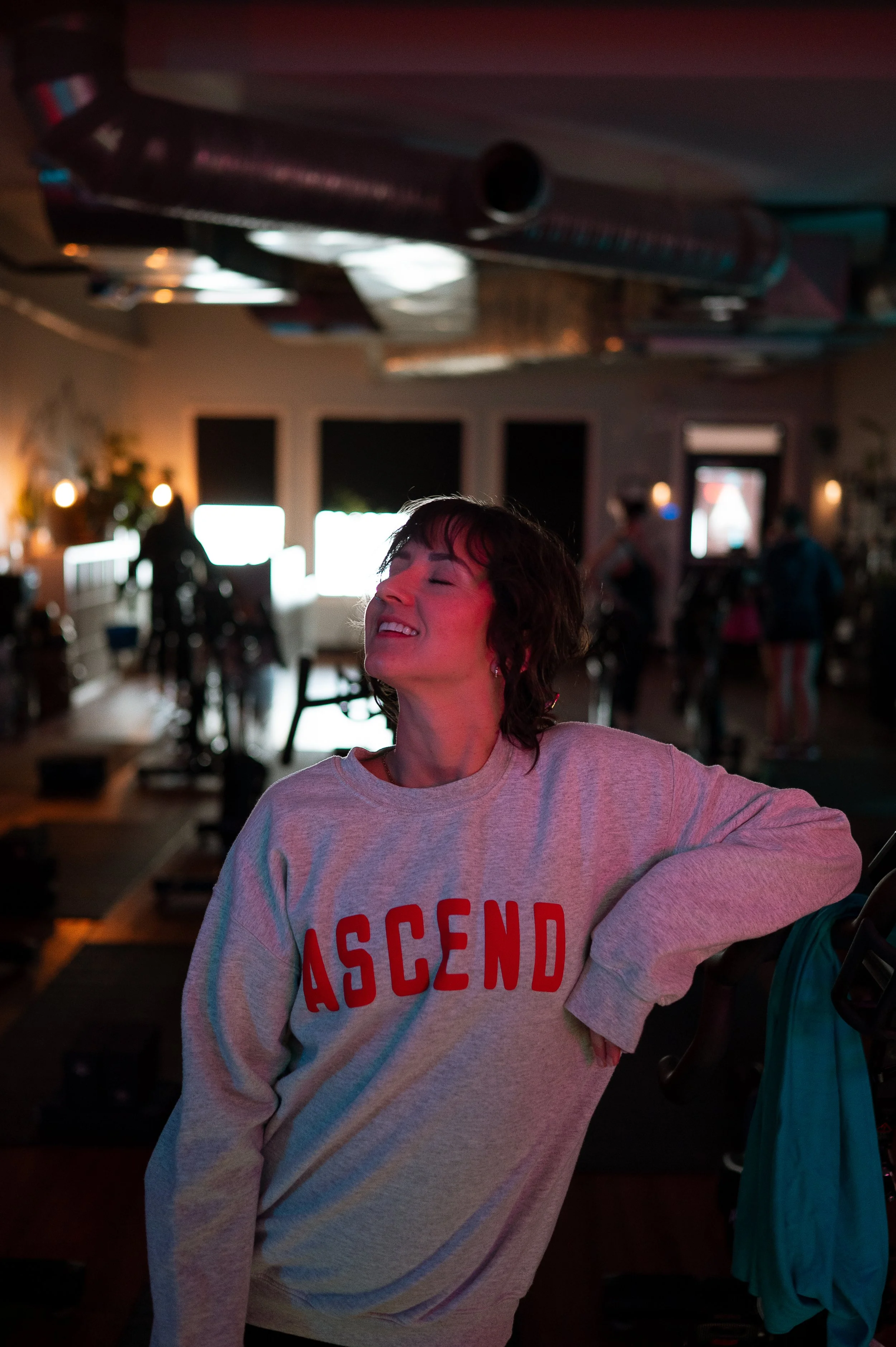 A woman with short dark hair smiling with closed eyes, wearing a light-colored sweatshirt that has red text reading 'ASCEND'. She is in a dimly lit indoor space with blurry figures and bright light sources in the background.
