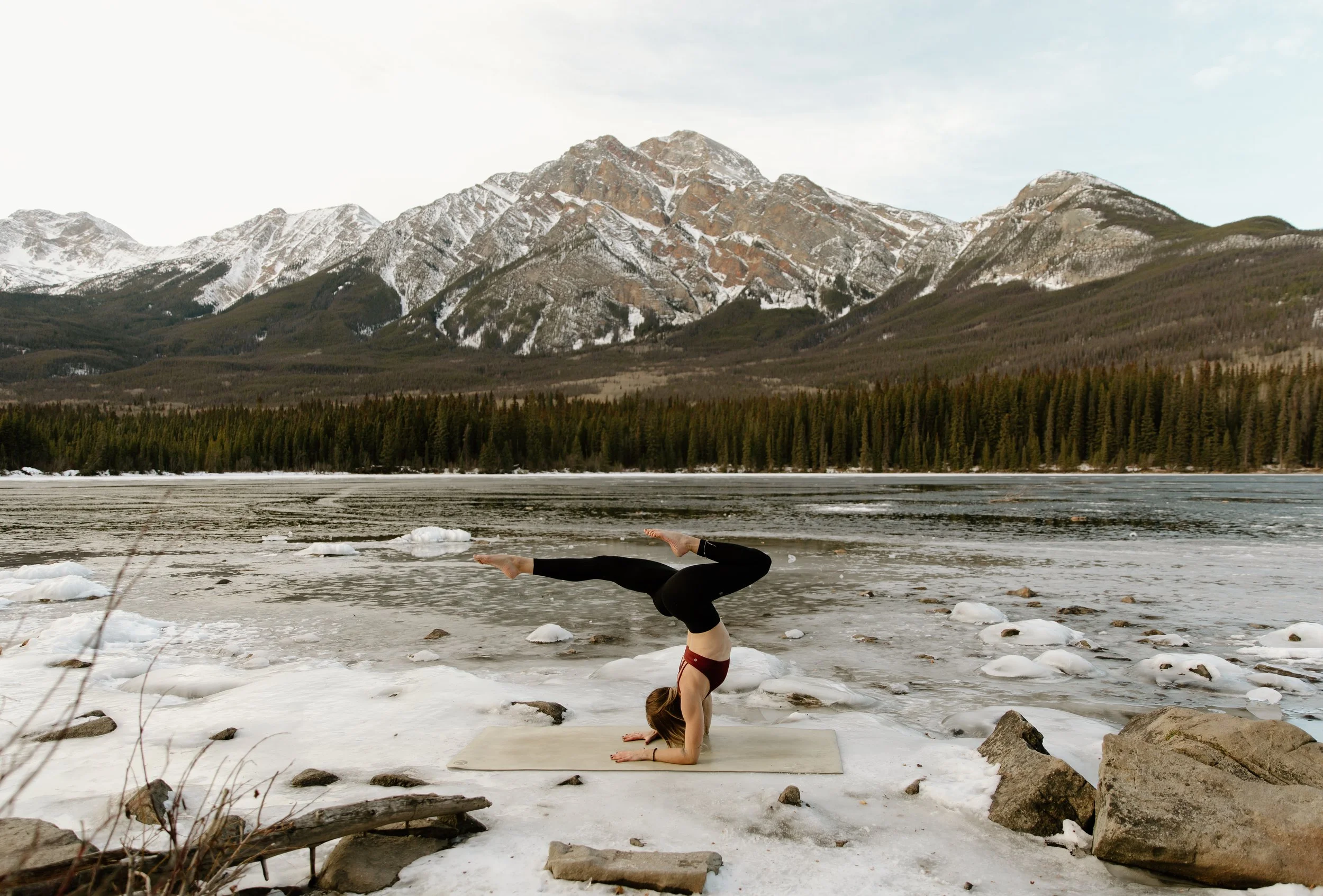 Person practicing yoga in the pose of headstand on a snowy outdoor area near a frozen lake with mountains and forest in the background.