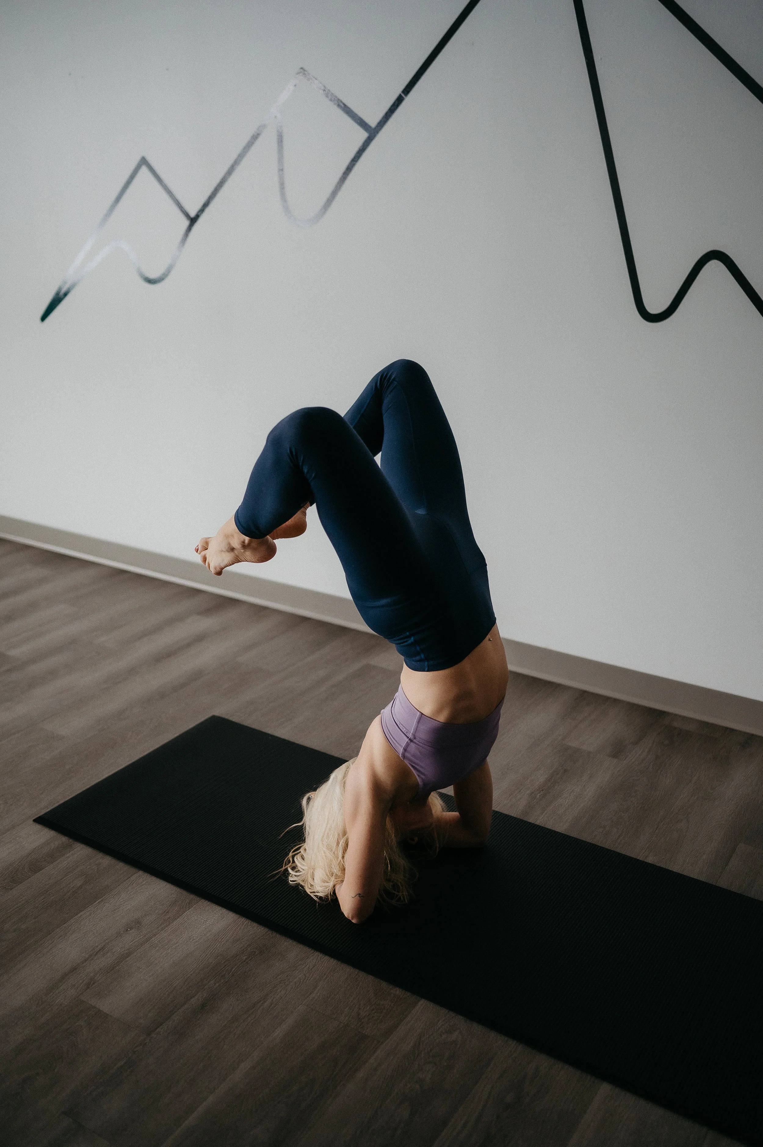 A woman with blonde hair practicing yoga indoors on a black mat, in a headstand pose, with a large painted line graph on the wall behind her.