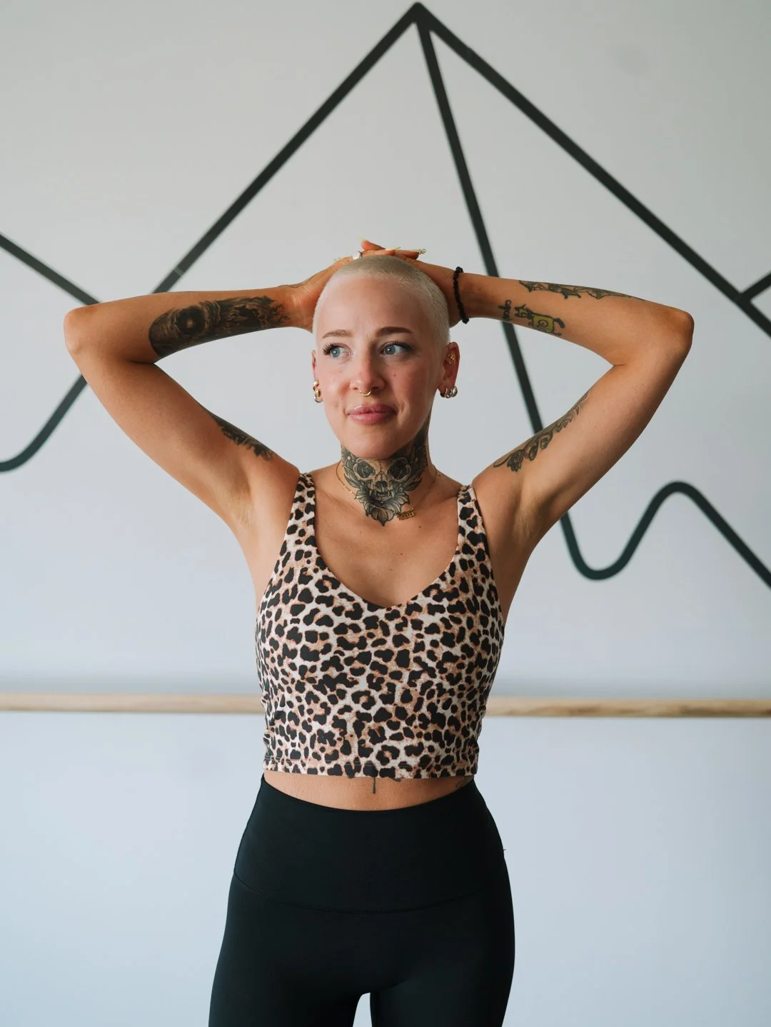 A woman with short blonde hair and tattoos on her arms, neck, and face, wearing a leopard print sports bra and black leggings, standing in a dance or fitness studio with a geometric wall design.