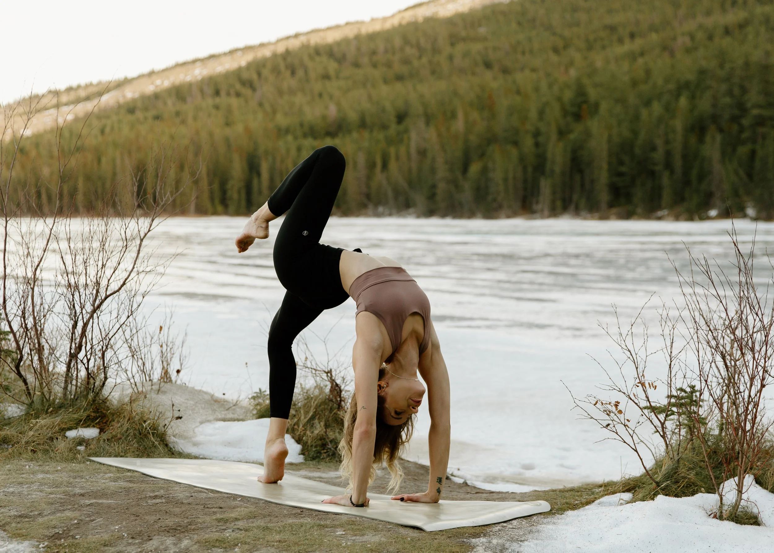 A woman practicing yoga outdoors near a body of water, performing a handstand on a yoga mat with a mountain and forest in the background