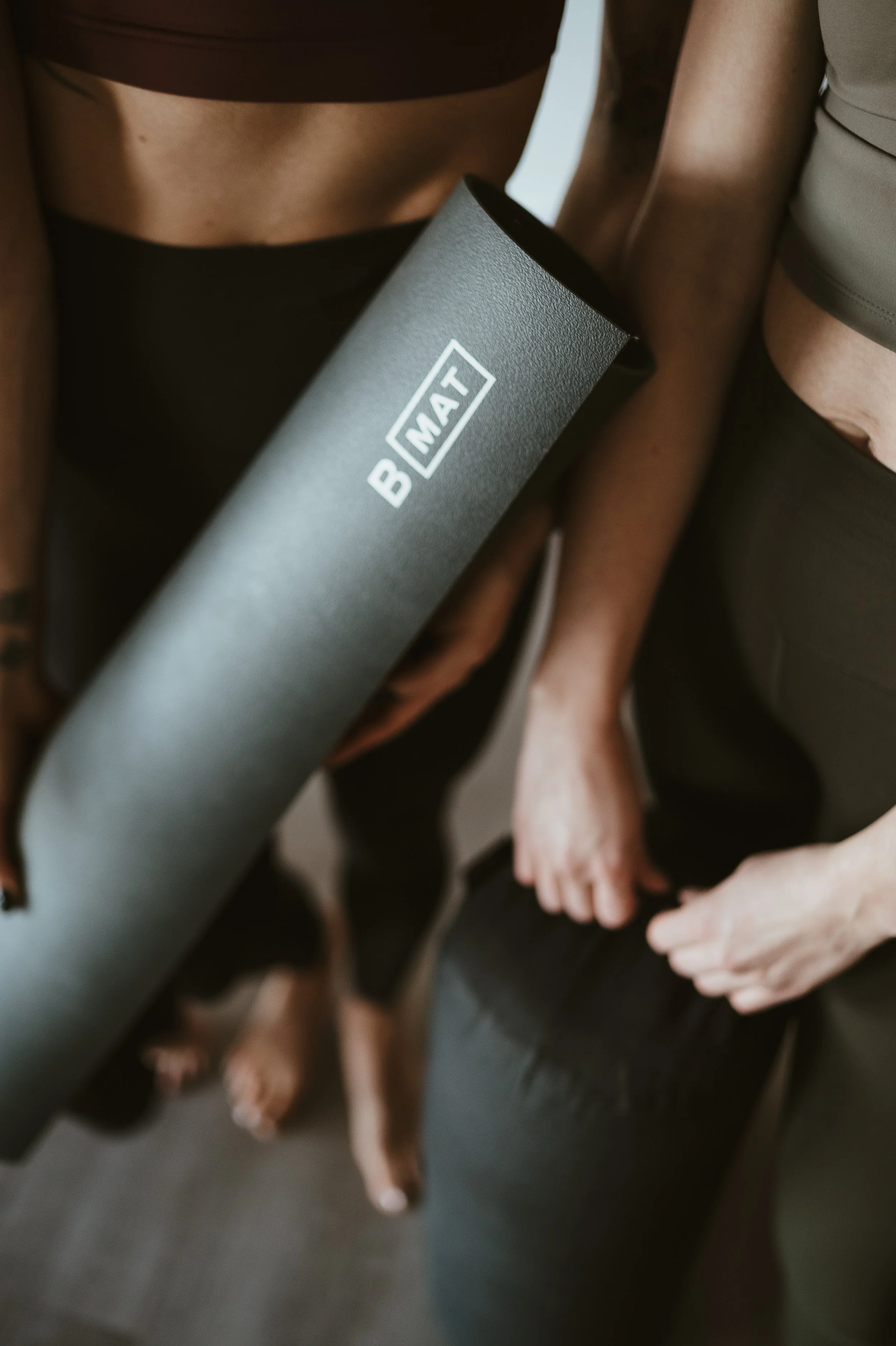 A person in workout attire holding a black yoga mat in front of them, with other people in the background.