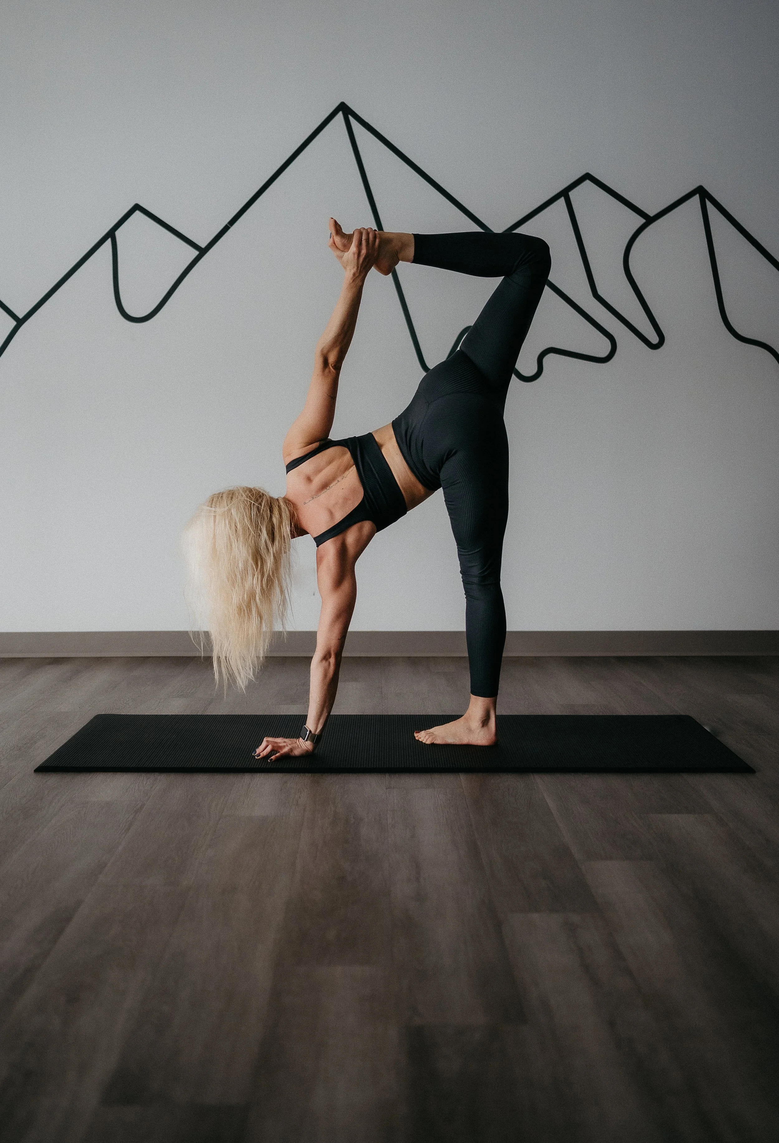 Woman practicing yoga in halfmoon pose on a black mat, in front of a wall with black mountain line art.