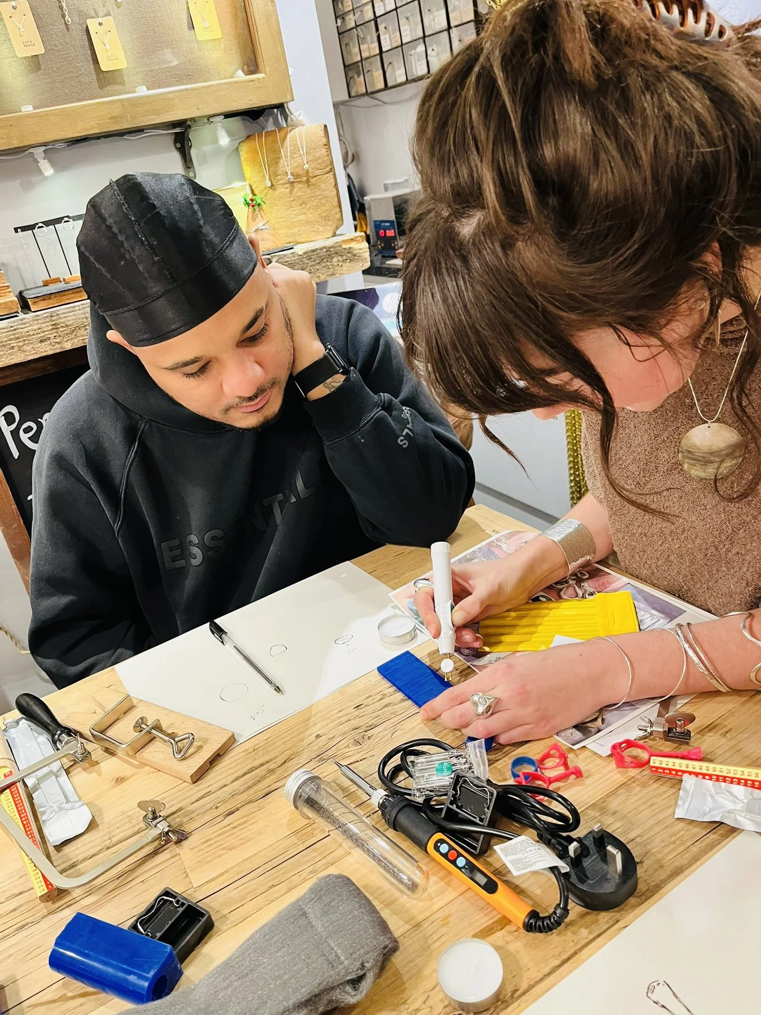 Two women working together at a wooden table filled with scientific and craft supplies, including a syringe, measuring tools, and measuring tape, in a workshop or creative space.