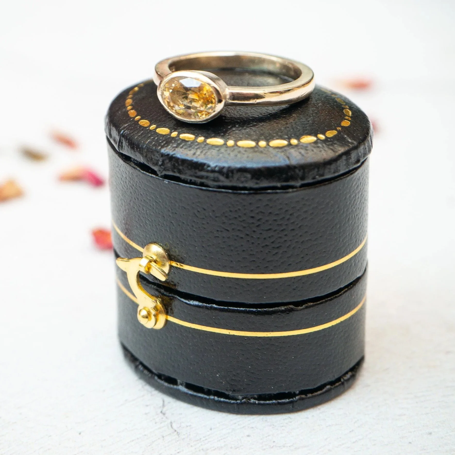 Close-up of a silver ring with a yellow gemstone, displayed on a black padded holder with gold accents. The background features scattered red, yellow, and white beads.