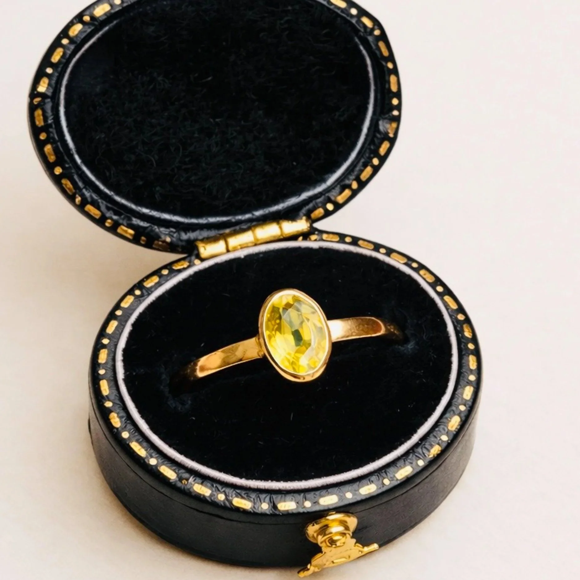 A black jewelry box with gold decorative accents, open to reveal a gold ring with a large yellow gemstone inside.