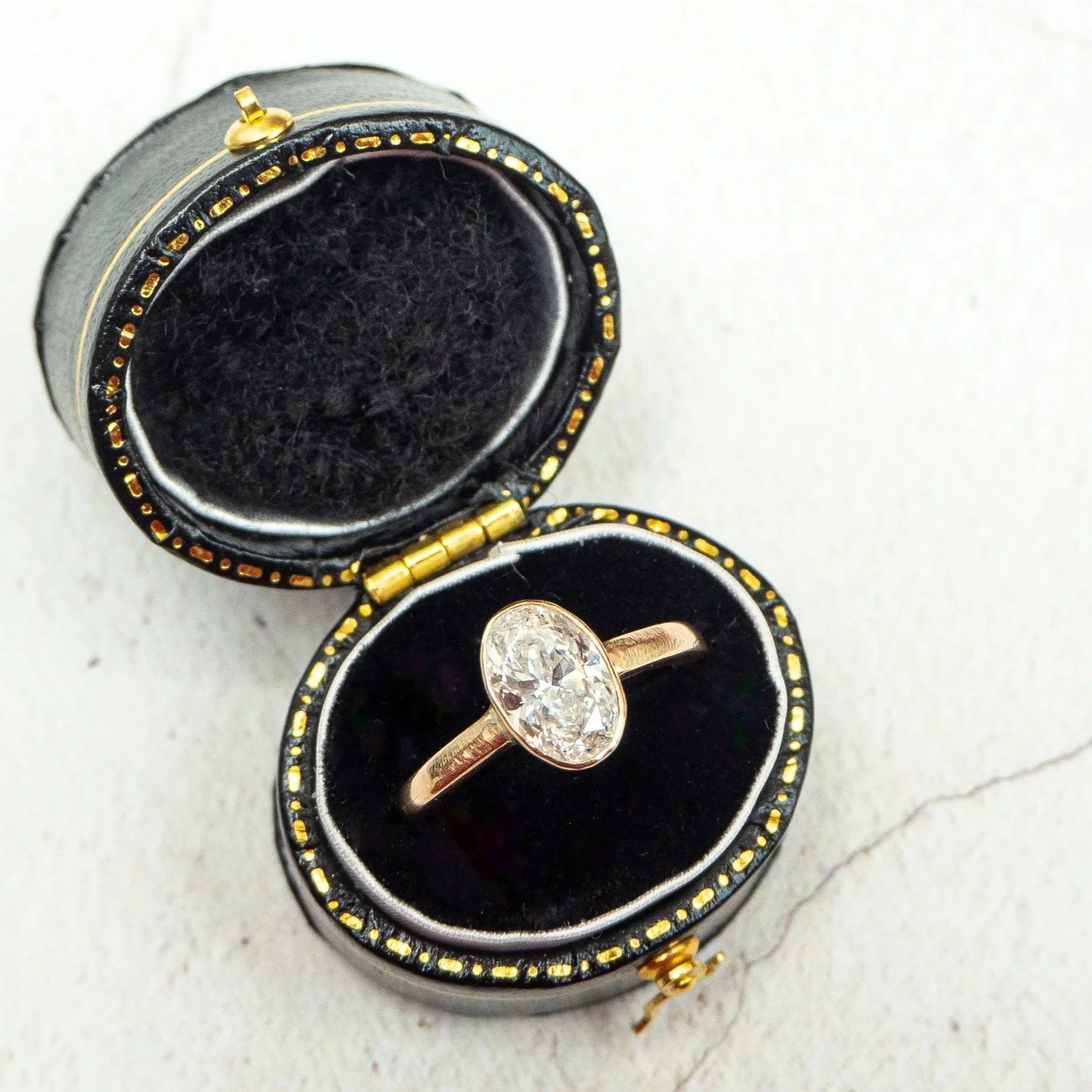 Open black jewelry box with gold trim containing a gold ring with a large clear gemstone, placed on a white textured surface.