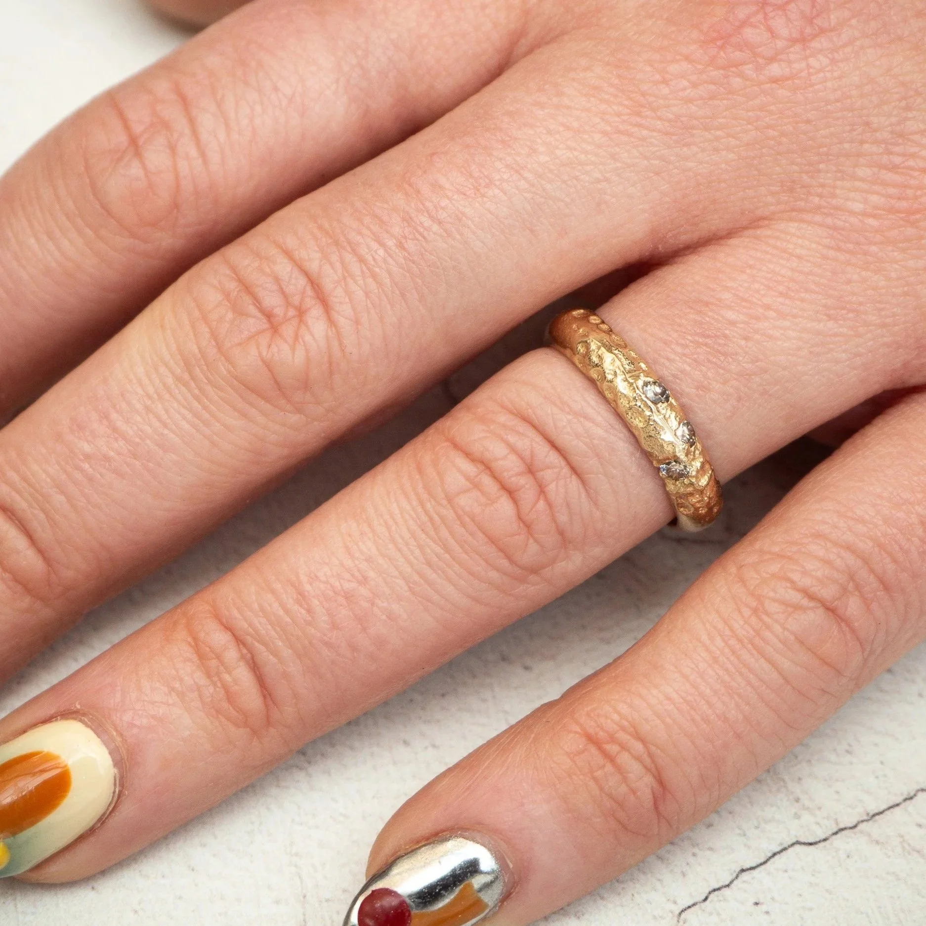 Close-up of a person's hand with multi-colored and textured nail polish on fingernails, and a textured gold ring with embedded stones on the ring finger.