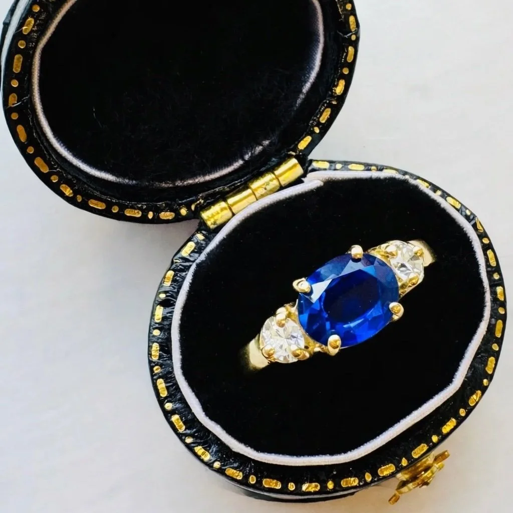 A gold ring with a large oval blue gemstone flanked by smaller clear gemstones, placed inside a black velvet jewelry box with gold accents.