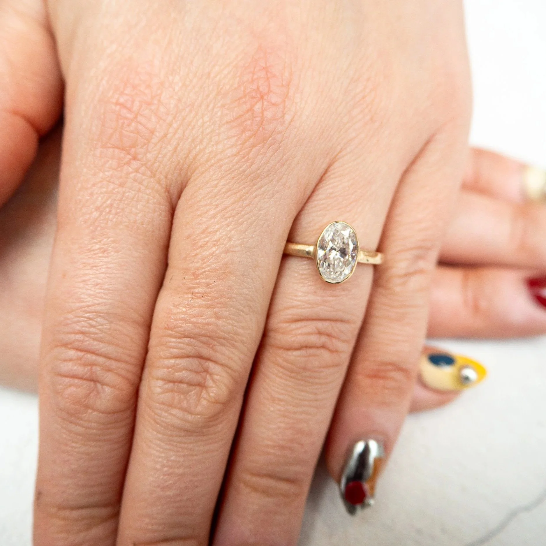 Close-up of a hand with a gold ring featuring a large oval diamond. The hand has light skin and short nails, with other rings visible on the fingers. The background includes a white surface and colorful objects.