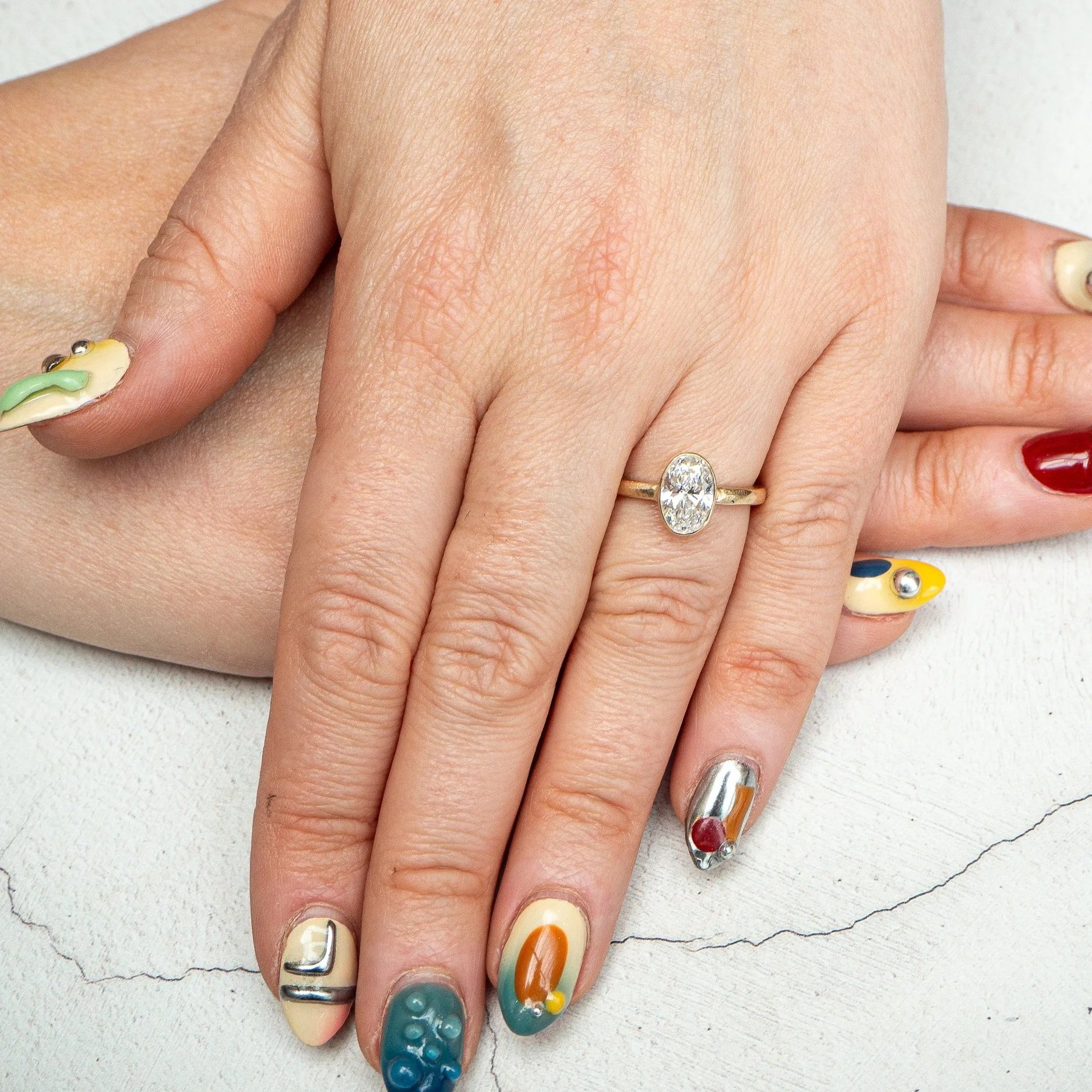 Close-up of a person's hand with colorful, decorative nail art. The person is wearing a diamond engagement ring. The background is a white surface with a curved line.
