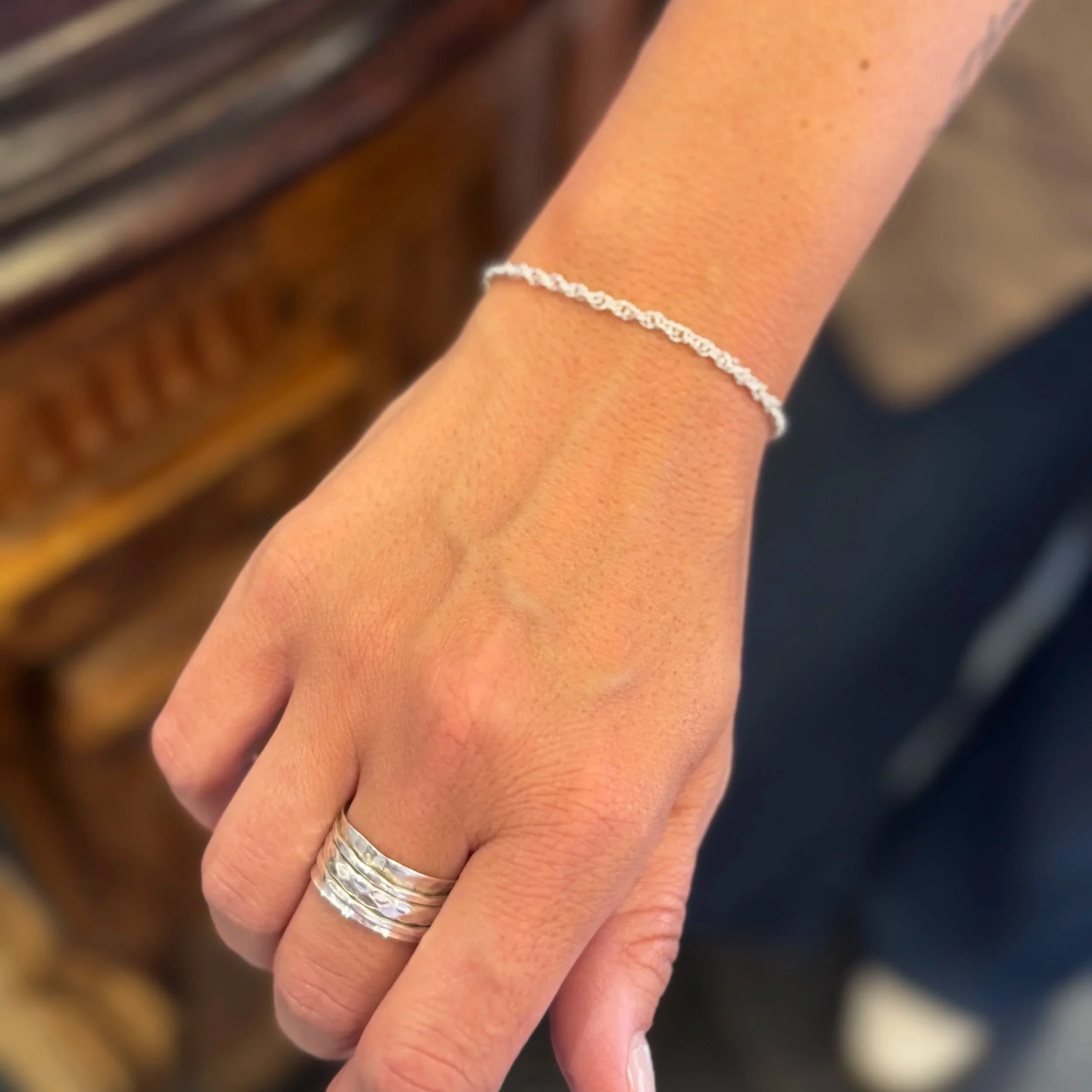 A close-up of a person's hand with a silver ring on the ring finger and a delicate silver bracelet on the wrist, with a blurred wooden surface in the background.