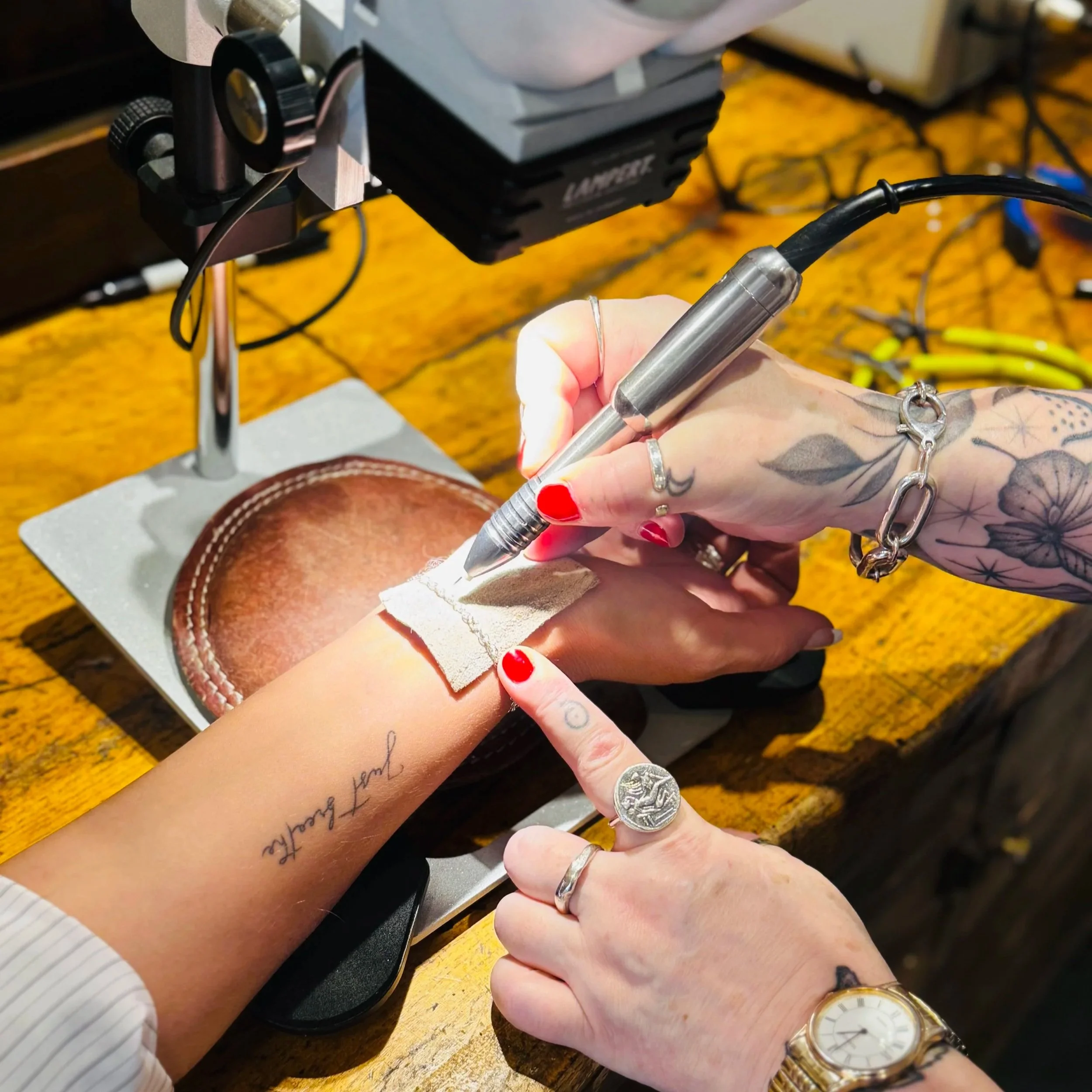 Tattoo artist using a tattoo machine on a person's wrist with a bandage, while the tattoo artist's hand is pointing at the wrist. The artist has tattoos, and wears rings and a watch. The workspace has tools and equipment for tattooing.
