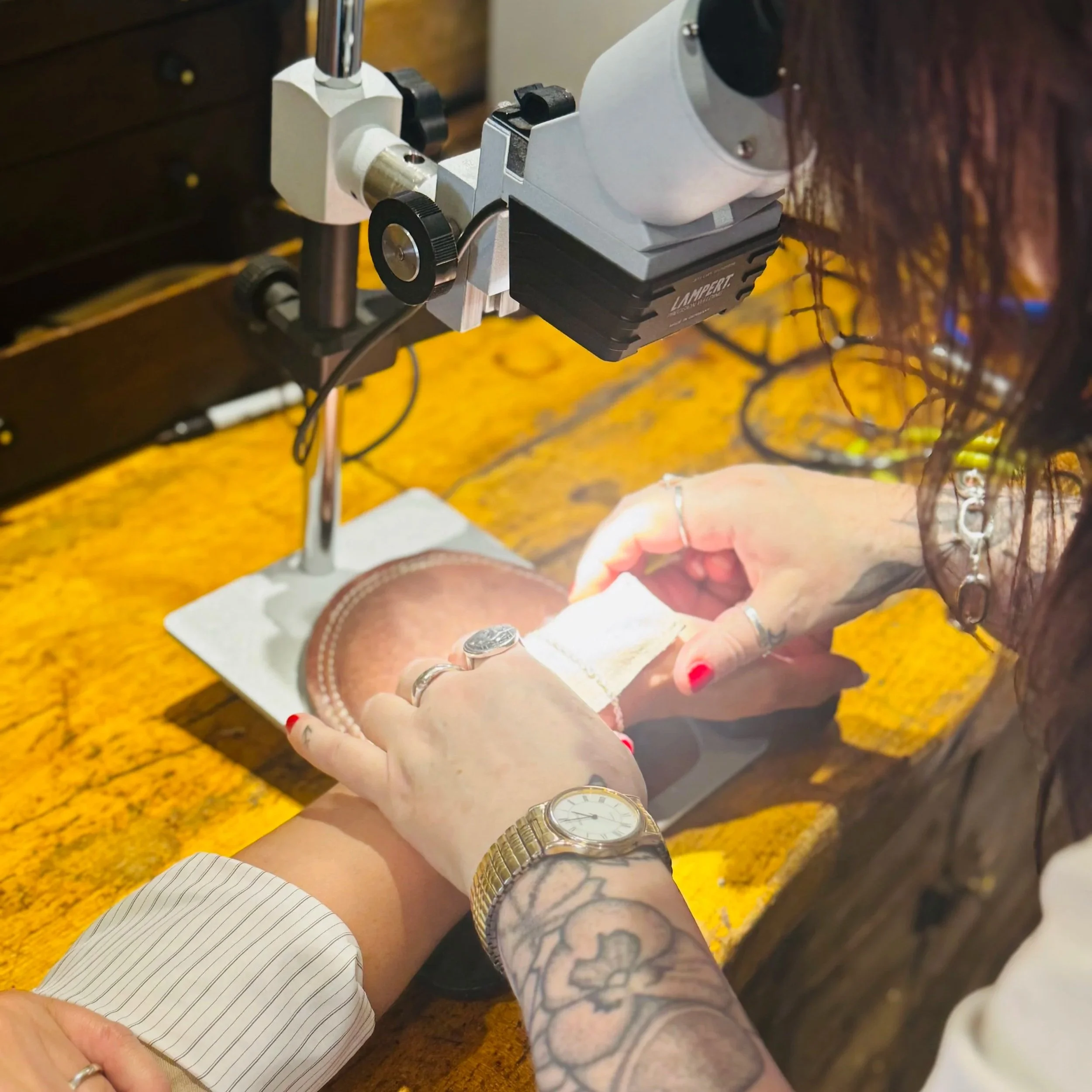 A person with tattoos on their arm and wearing a gold watch is operating a watchmaker's loupe to examine a leather wallet on a wooden workbench, with another person holding the wallet steady.