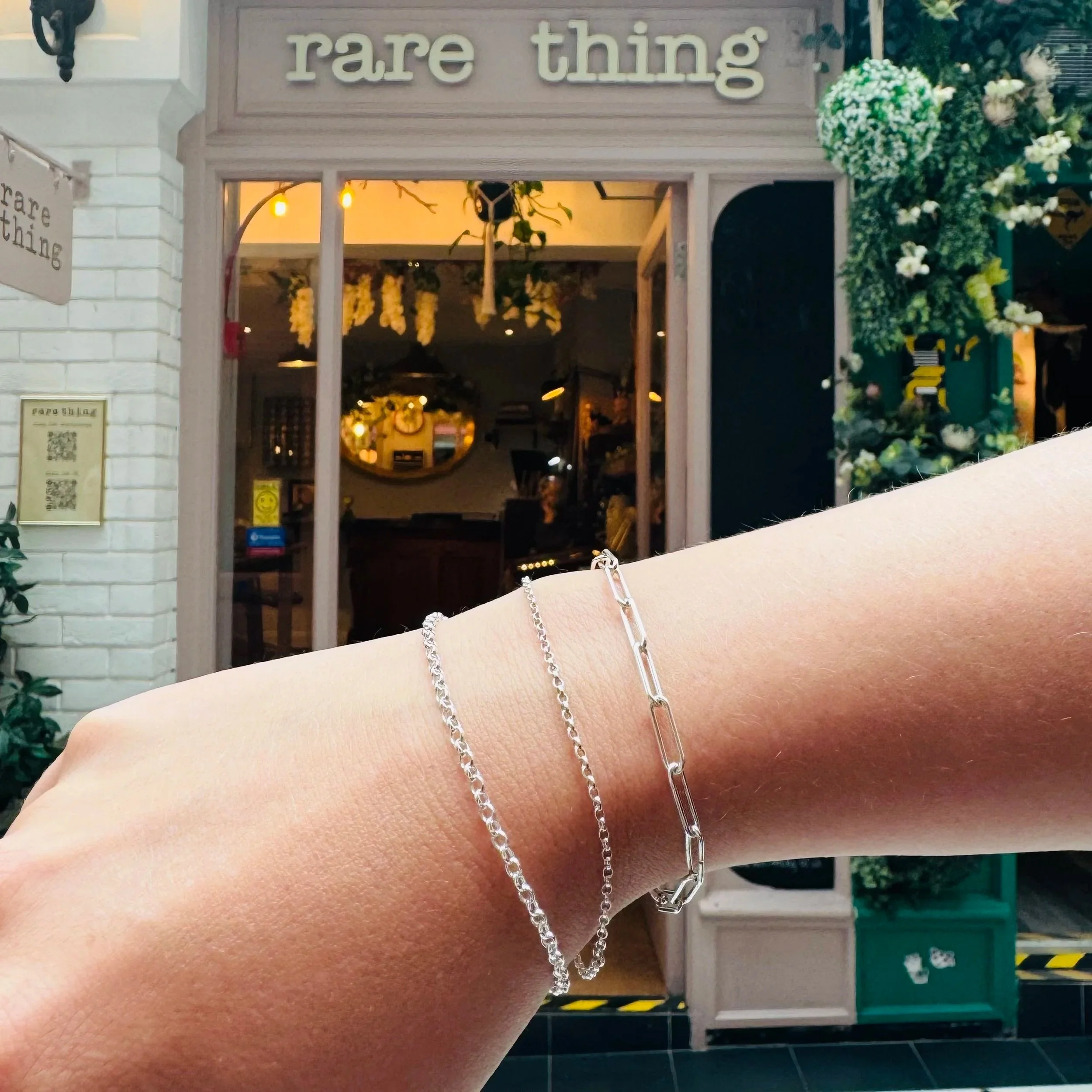A close-up of a person's wrist wearing three silver bracelets in front of a shop entrance with a "rare thing" sign. The shop is decorated with flowers and plants.