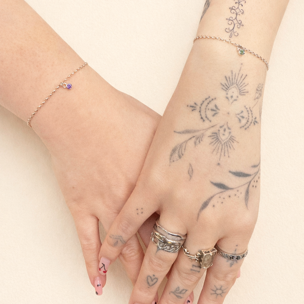 Two hands with various tattoos and multiple rings on the fingers, and wearing delicate bracelets, are intertwined against a light background.