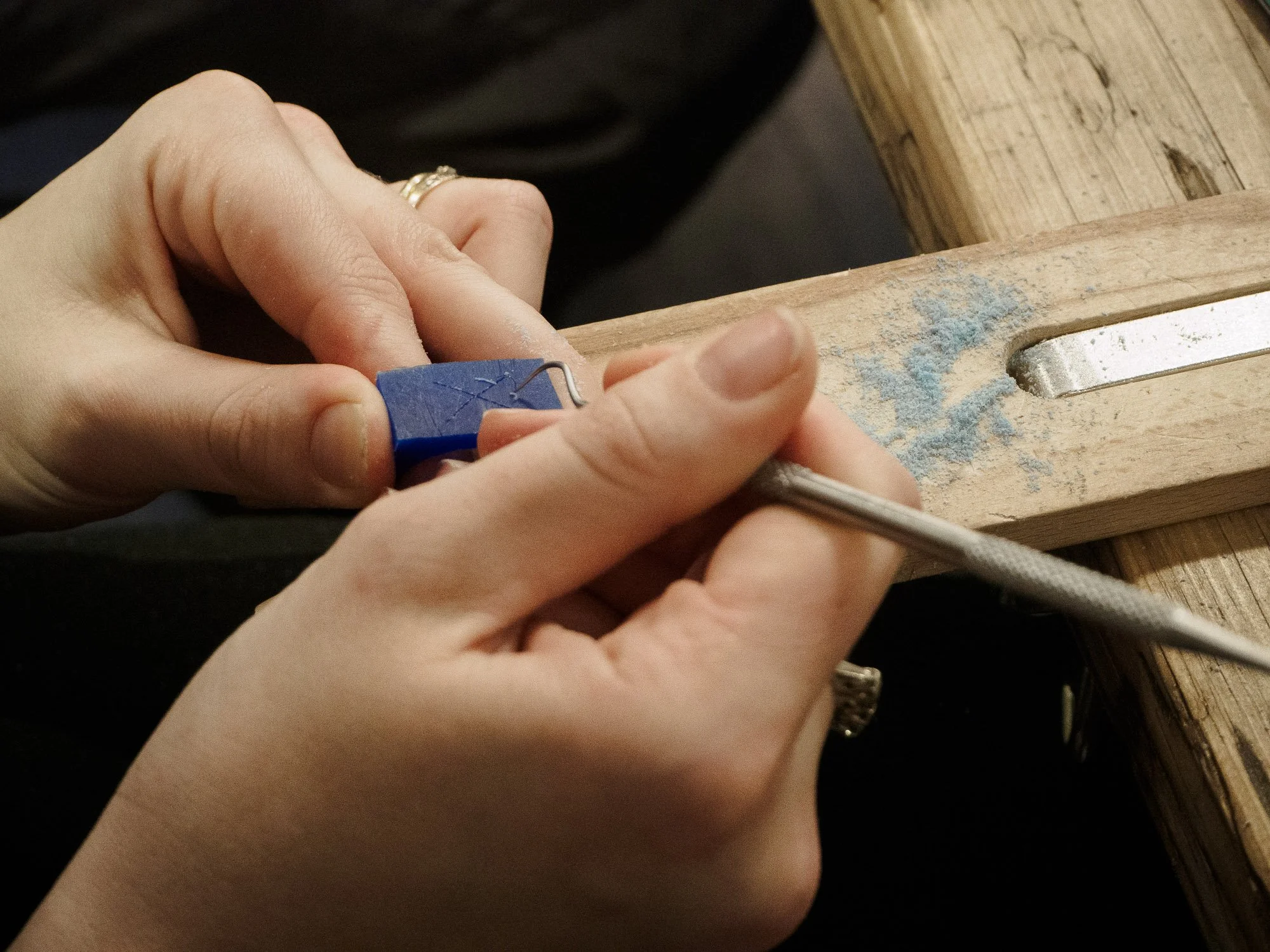 A person using a dental pick and a blue tool to sand or carve a small piece of material, with wood shavings around and a wooden work surface.