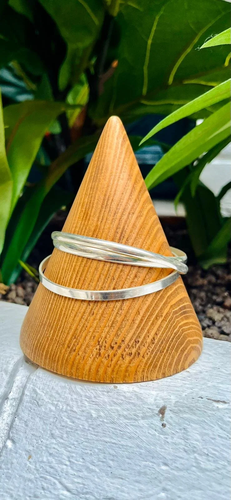 Two silver bangles perched on a wooden cone, with green leaves in the background.