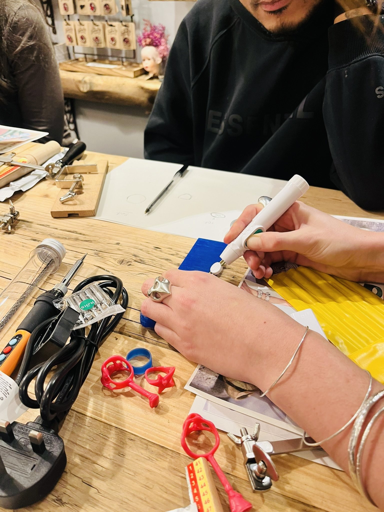 Person drawing on a small blue object with a white marker, surrounded by tools and colorful plastic rings on a wooden table.