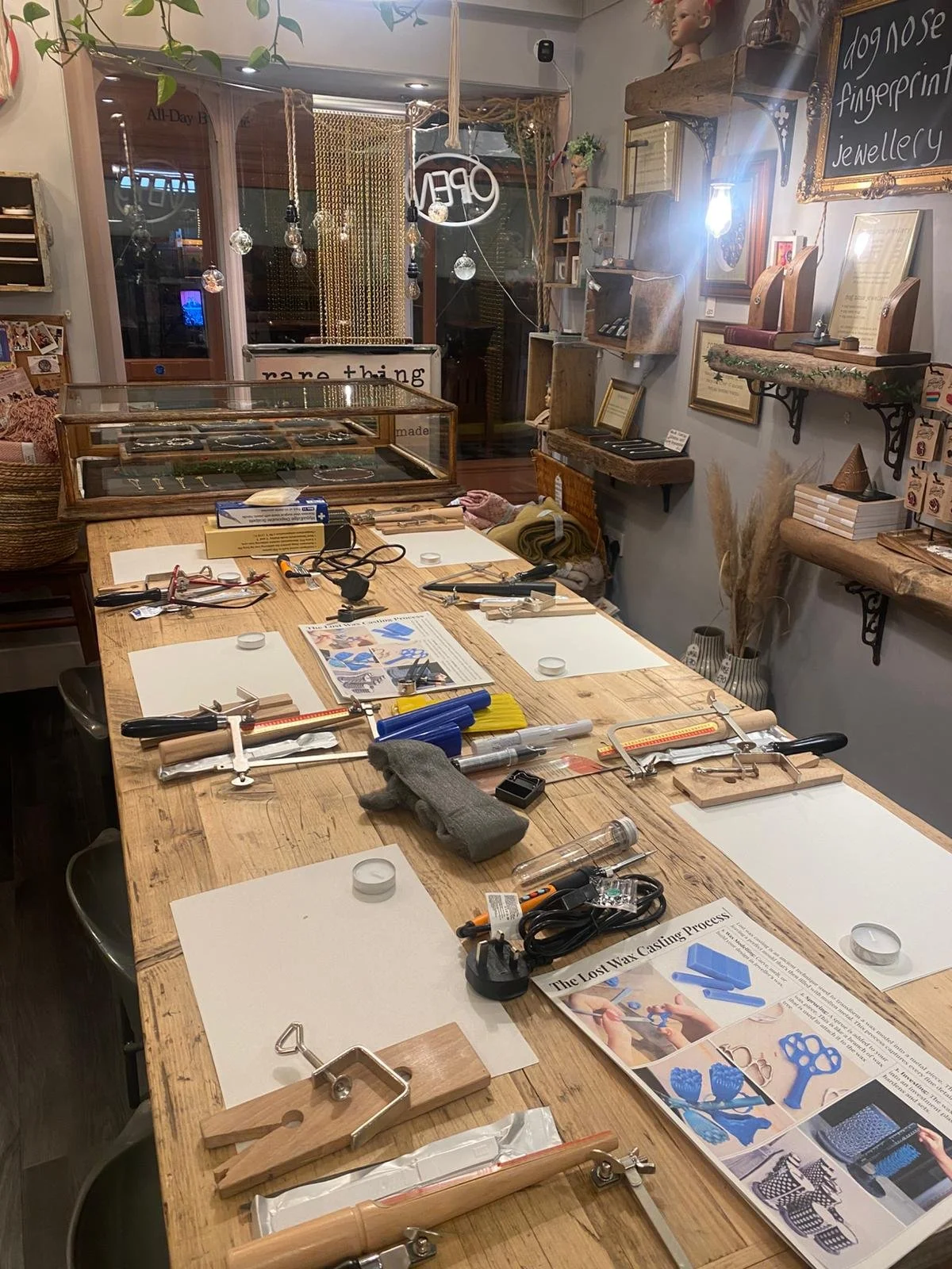 A craft workshop with a large wooden table set with various tools and supplies, including a metal roller, clamps, and papers, inside a cozy store with shelves displaying framed art, jewelry, and decor items.