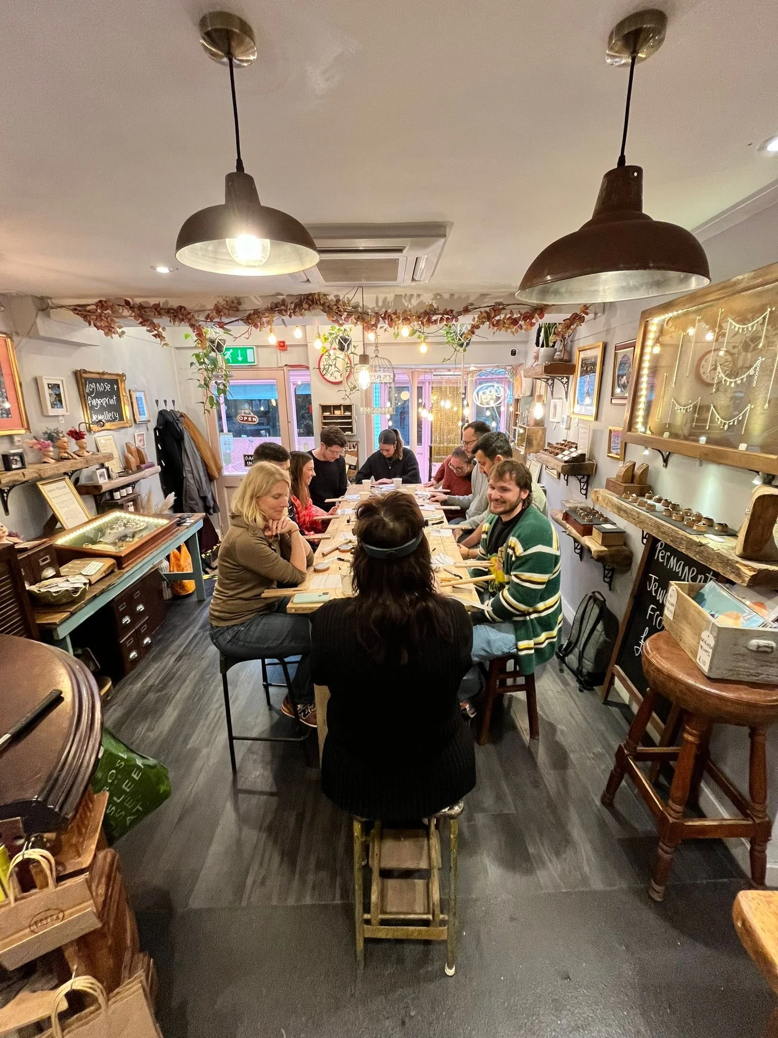 People sitting around a long wooden table in a cozy, decorated cafe during evening, engaging in conversation and activities.