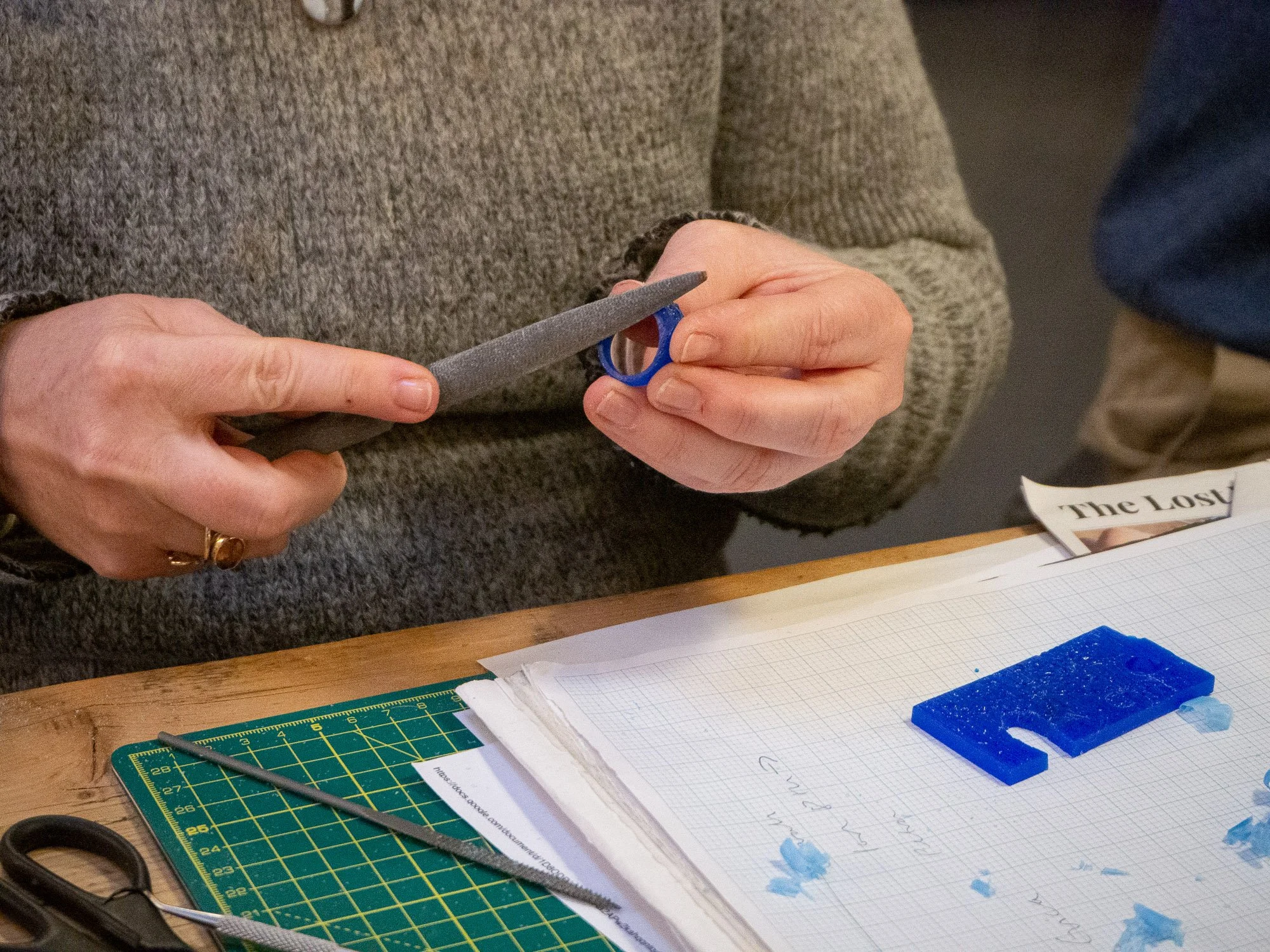 Person holding a small hand saw and a piece of blue material, possibly a plastic or resin, working on a craft project at a table with paper, tools, and cutting mat.