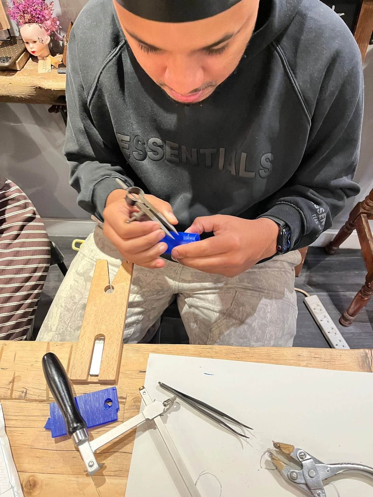A person wearing a black sweatshirt and a watch is sitting at a wooden table, working on a small blue object with a tool. The table has various tools, including pliers, tweezers, and a small clamp, as well as a piece of paper or foam pad. There is a 
