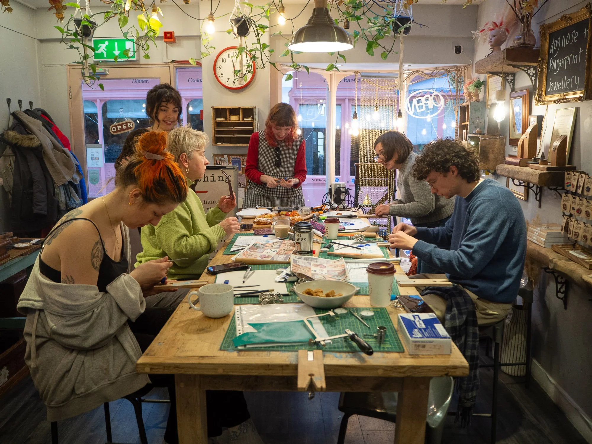 Group of six women gathered around a wooden table in a cozy craft shop, working on jewelry making projects with various tools and materials. The shop has decorative shelves, a clock, and windows with the word 'Open' visible outside, decorated with pl