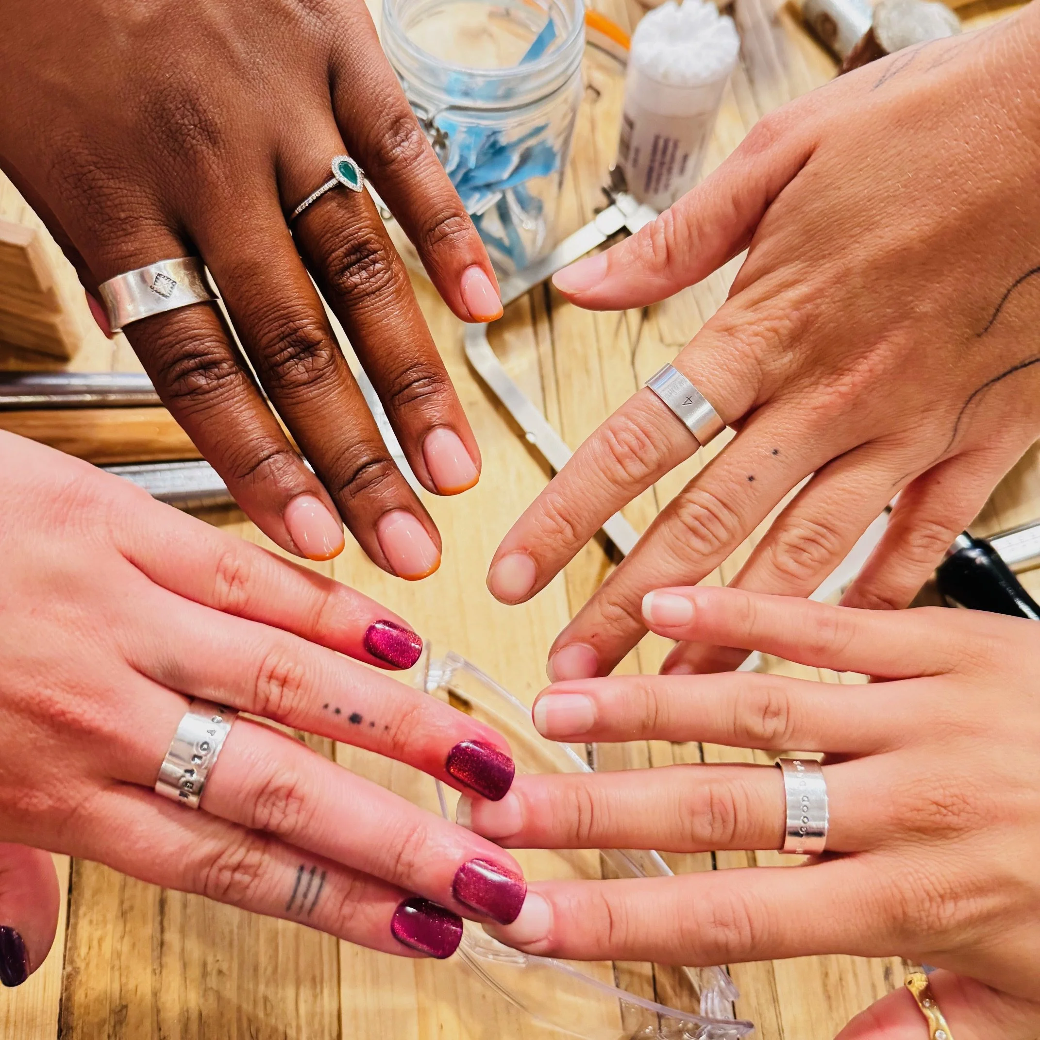 Four hands, three with rings, touching or arranging fingers, on a wooden surface with jewelry-making supplies nearby.
