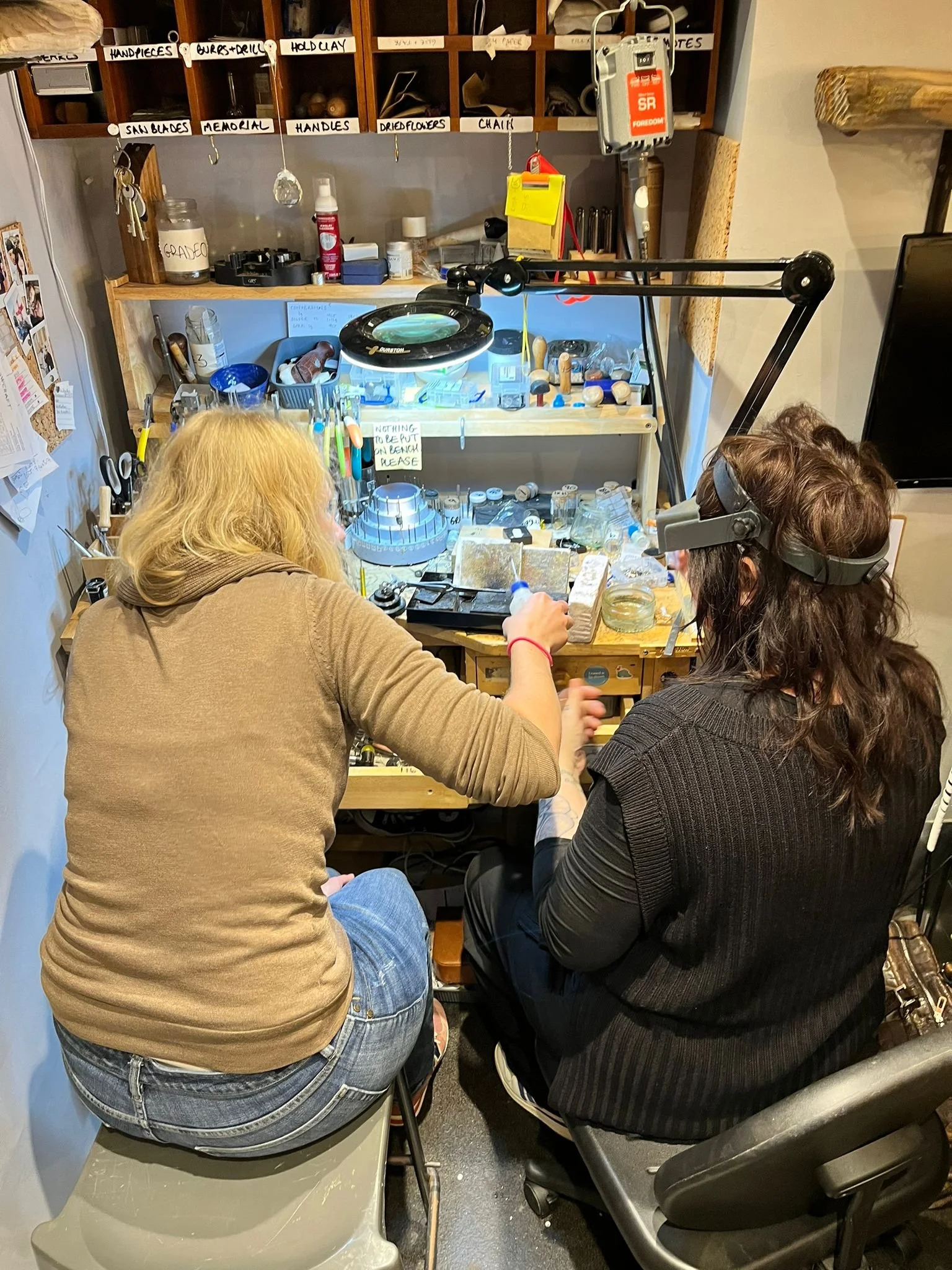 Two women working at a jewelry or craft workbench with tools, jars, and small objects, one using a magnifying headset, in a cluttered workshop with labeled wooden shelves and various supplies.