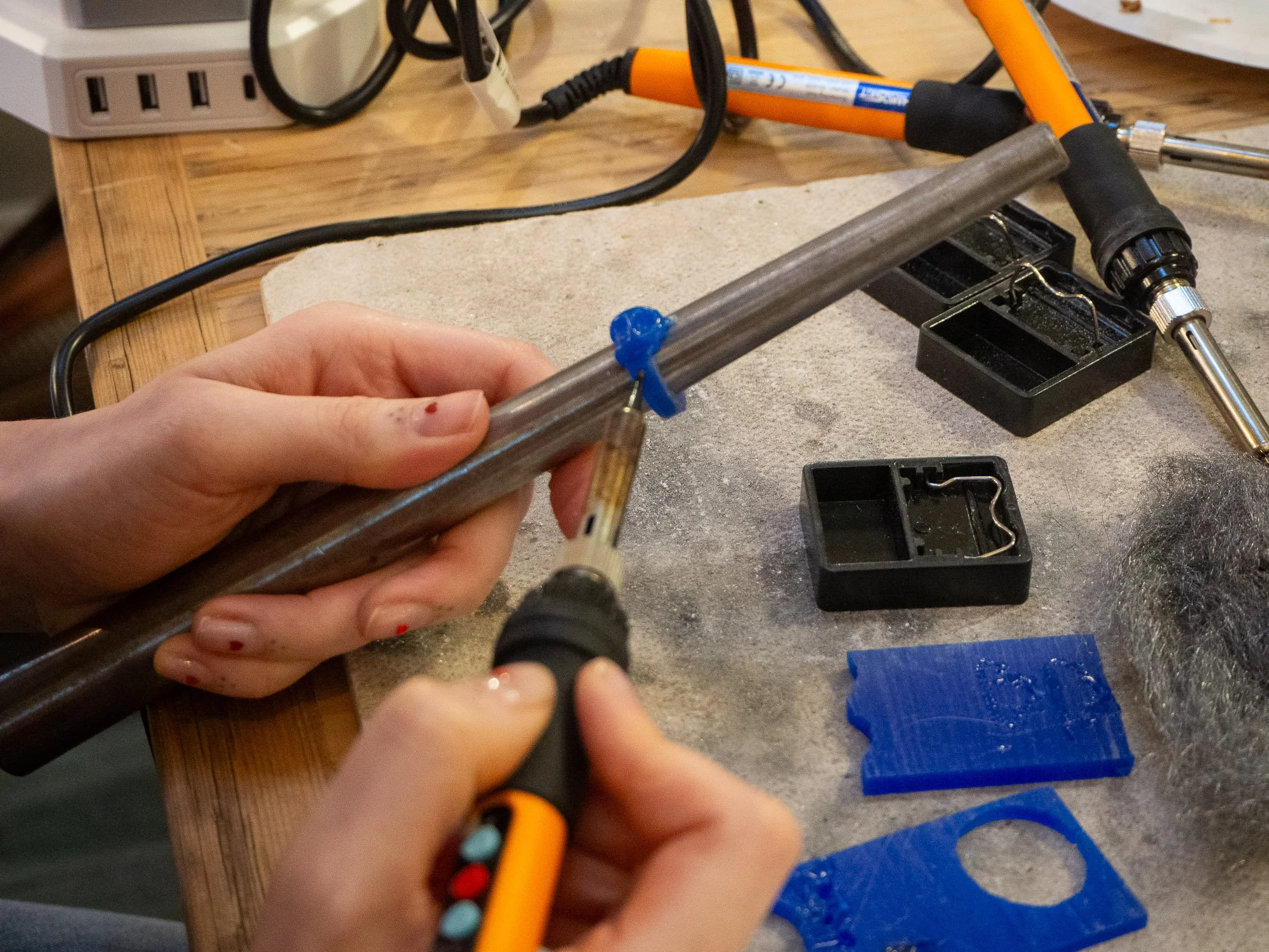Person soldering metal pipe with soldering iron at a workbench, surrounded by tools and small containers.