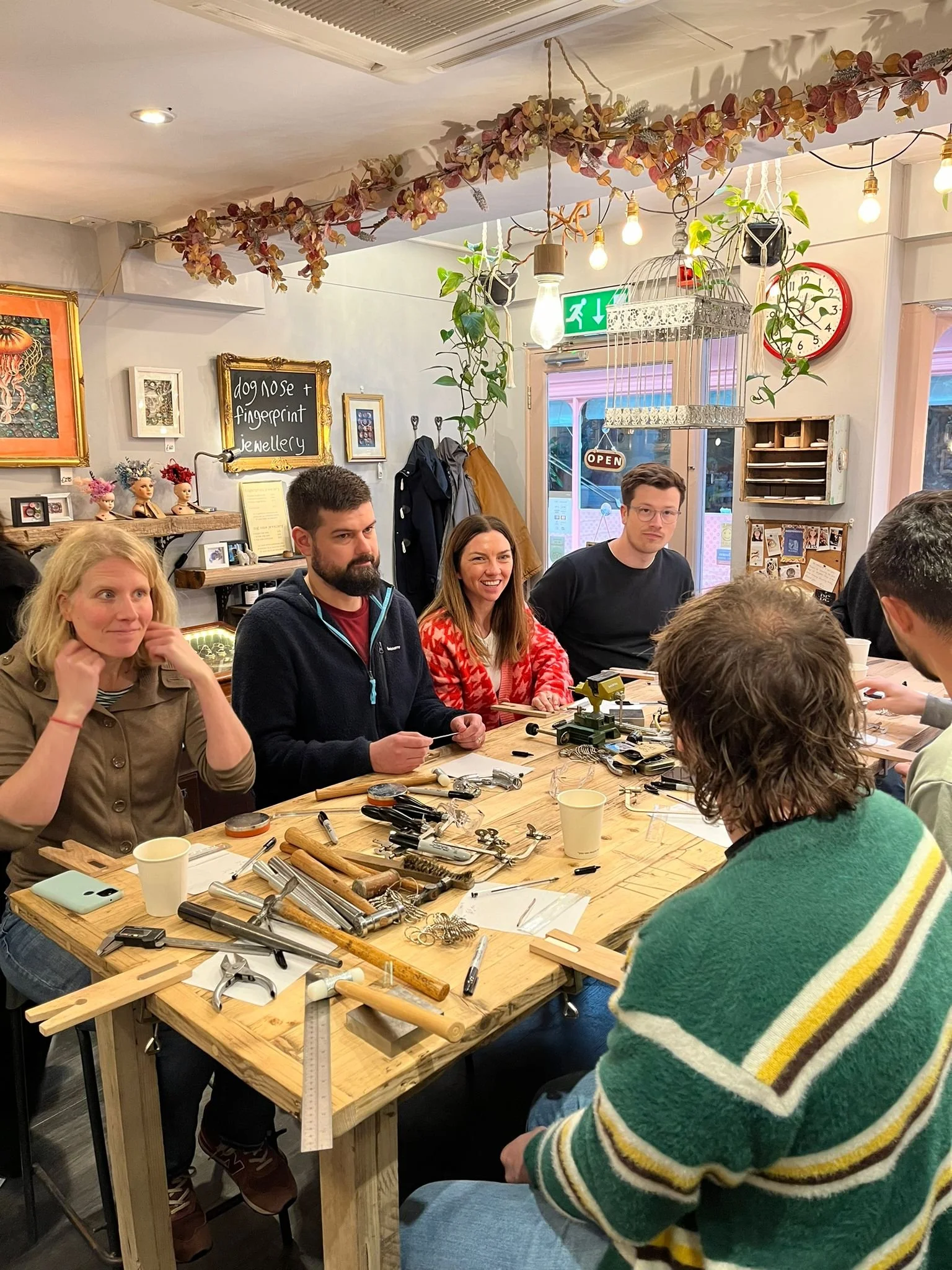 A group of people sitting around a wooden table with jewelry-making tools inside a cozy, decorated shop or workshop, with shelves, paintings, and plants in the background.