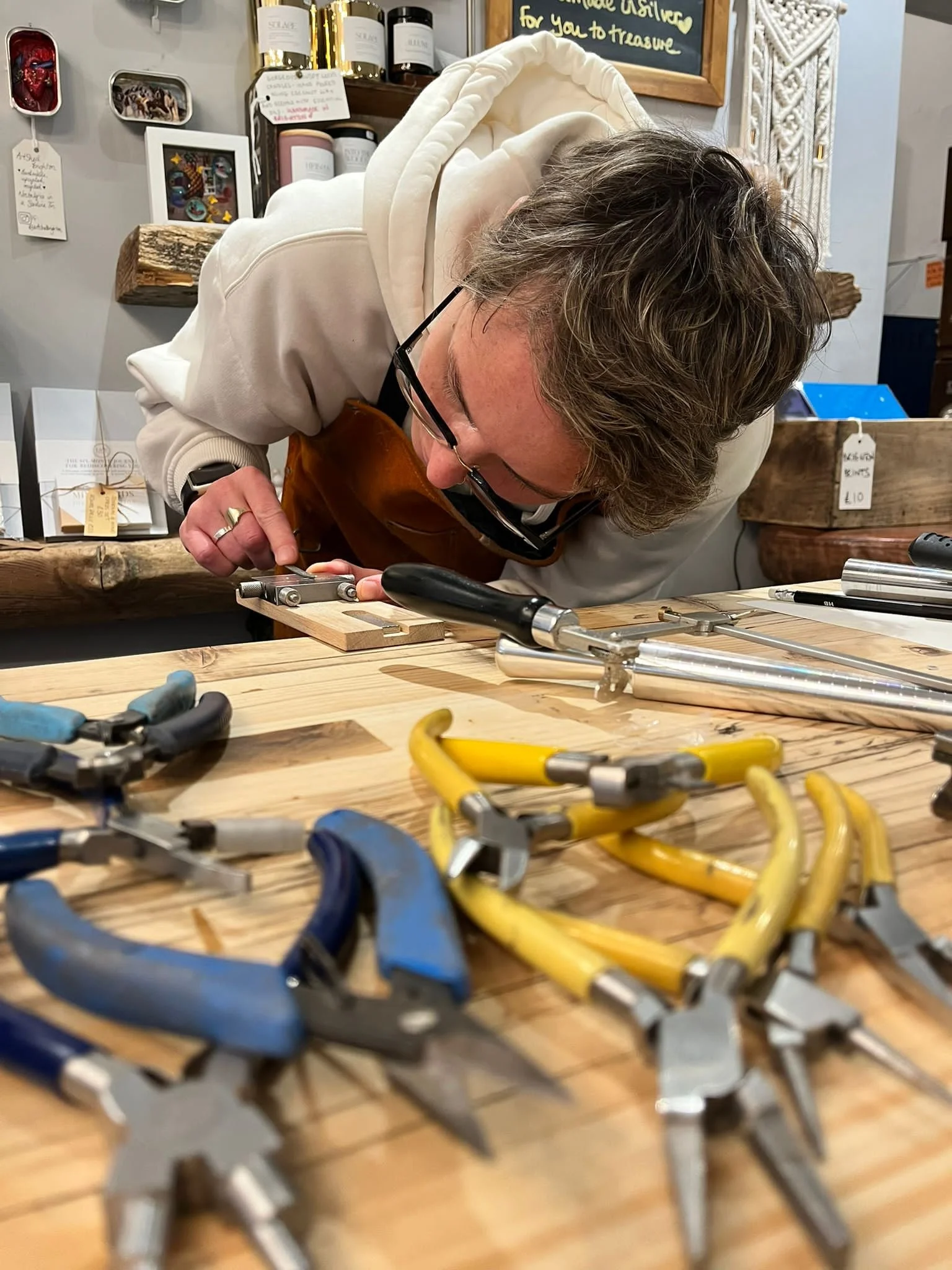 A person with glasses wearing a white hoodie and apron, working with woodworking tools on a wooden workbench in a workshop or craft store. Various clamps are scattered on the table, with shelves and framed pictures in the background.
