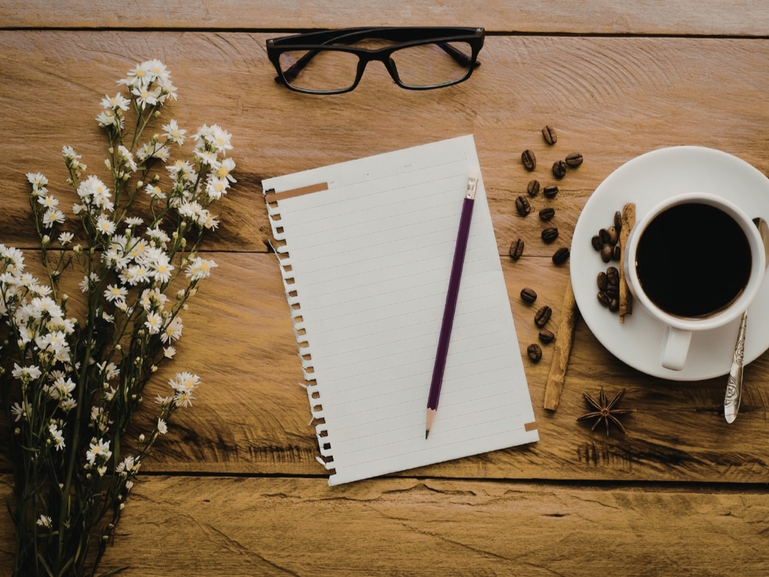 Notebook with a purple pencil, a pair of black glasses, a steaming cup of coffee on a saucer, scattered coffee beans, a cinnamon stick, and a star anise on a wooden surface with a bunch of daisies on the side.